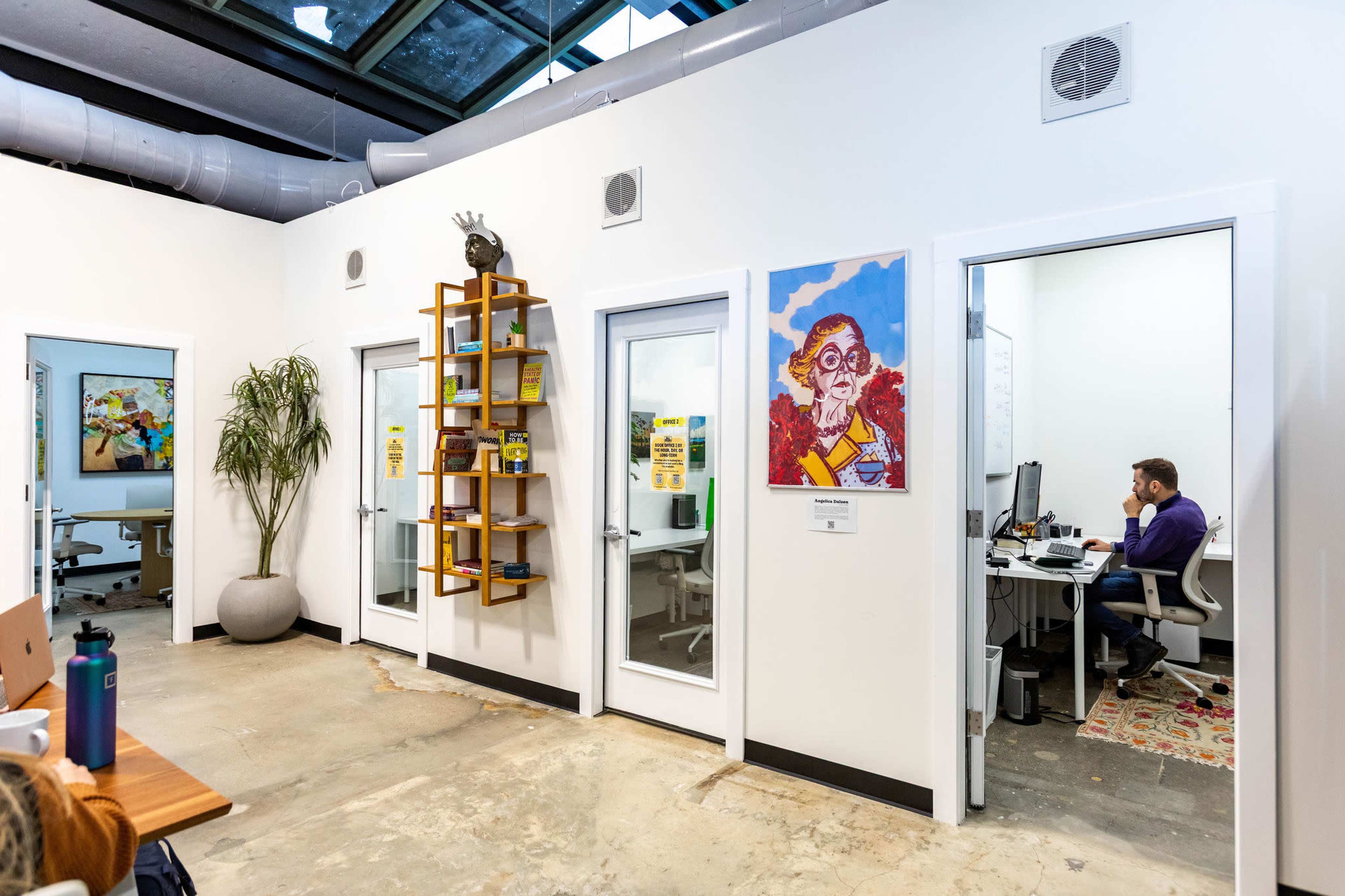The image shows a modern office space with white walls, glass doors, and a plant, featuring a decorative bookshelf and a person working at a desk in a separate room.