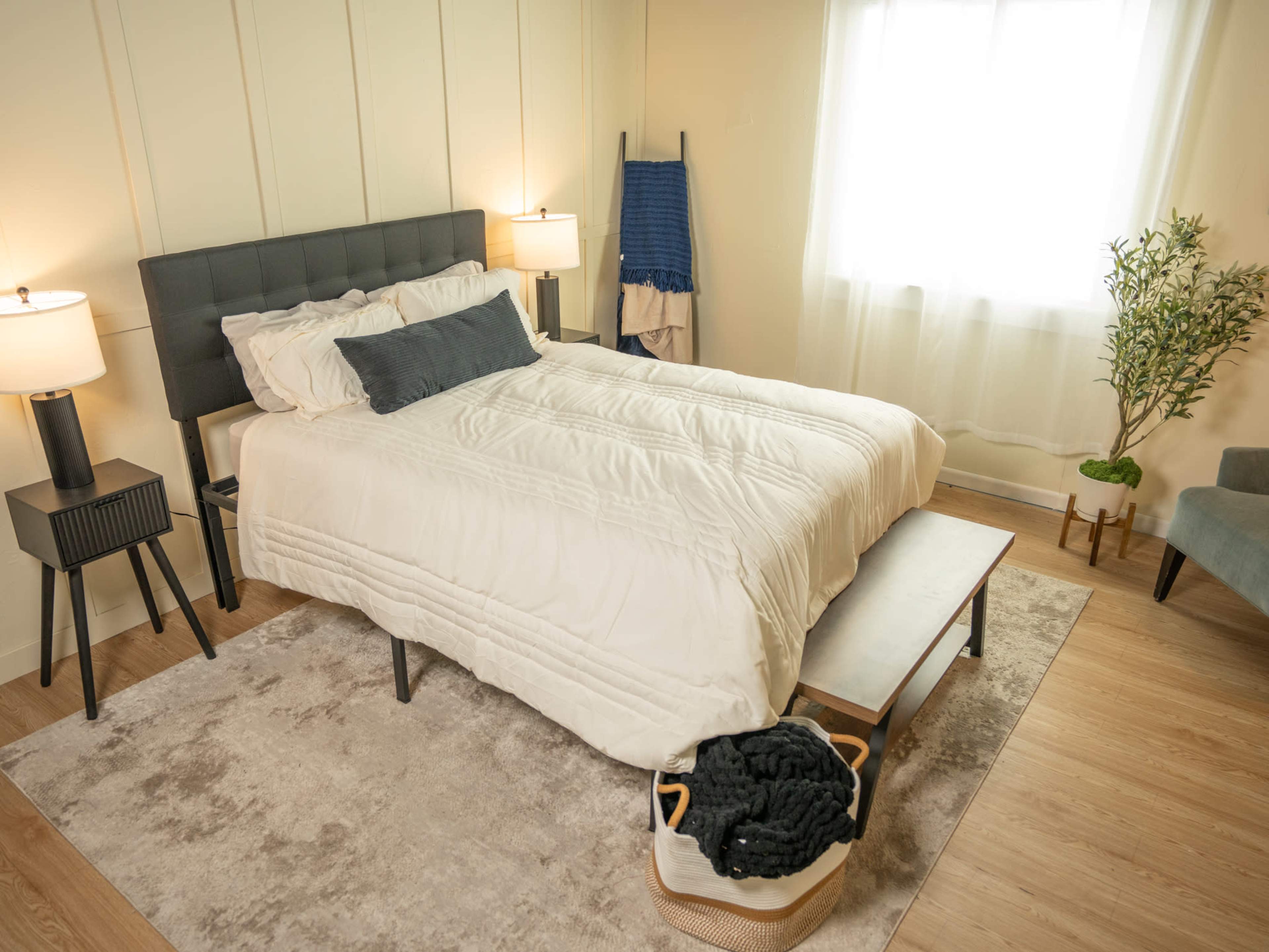 The image depicts a neatly arranged bedroom featuring a large bed with white linens, two bedside lamps, a small bench at the foot of the bed, and natural light streaming in from a nearby window.