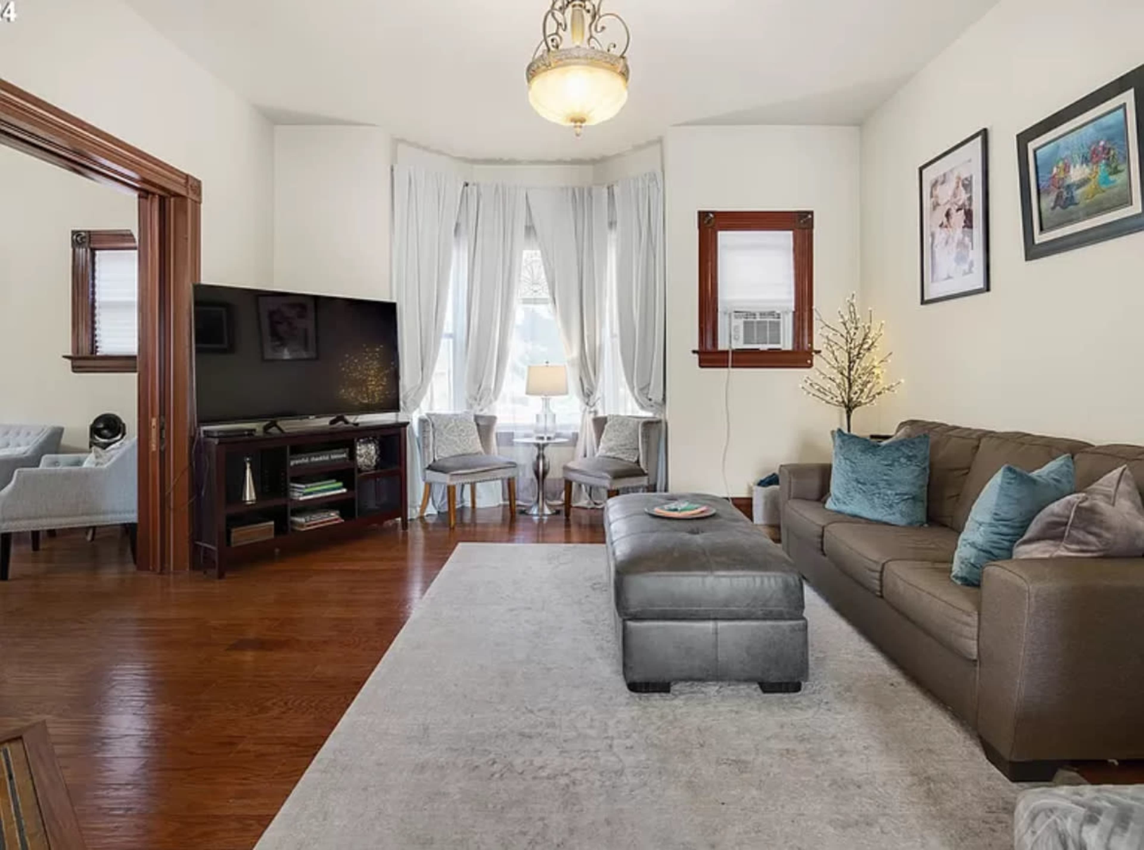 A living room features a gray sofa, an ottoman, a television, and two chairs near a window with sheer curtains.