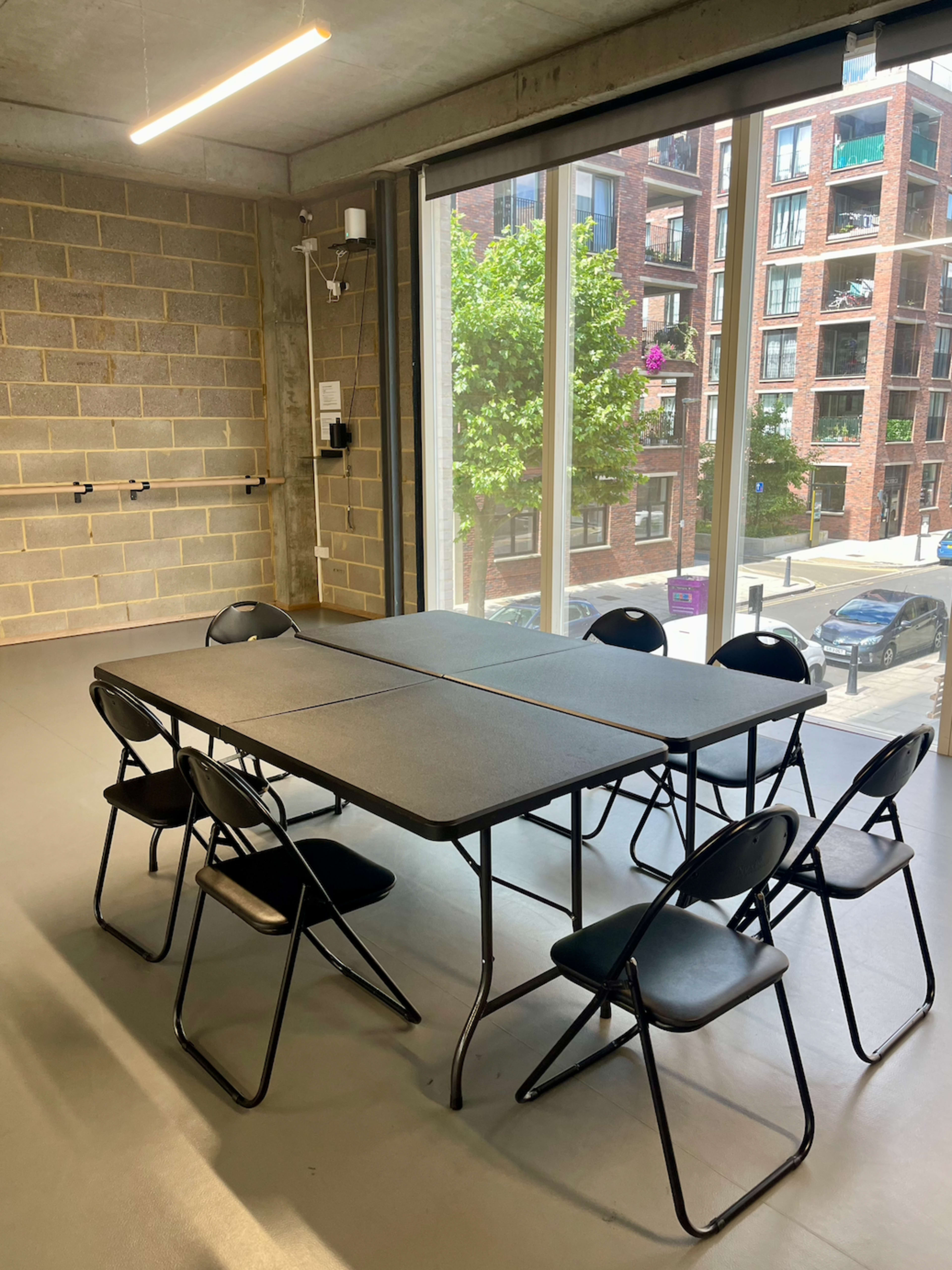 A rectangular table with black folding chairs is set up in a room with large windows overlooking a street and nearby buildings.