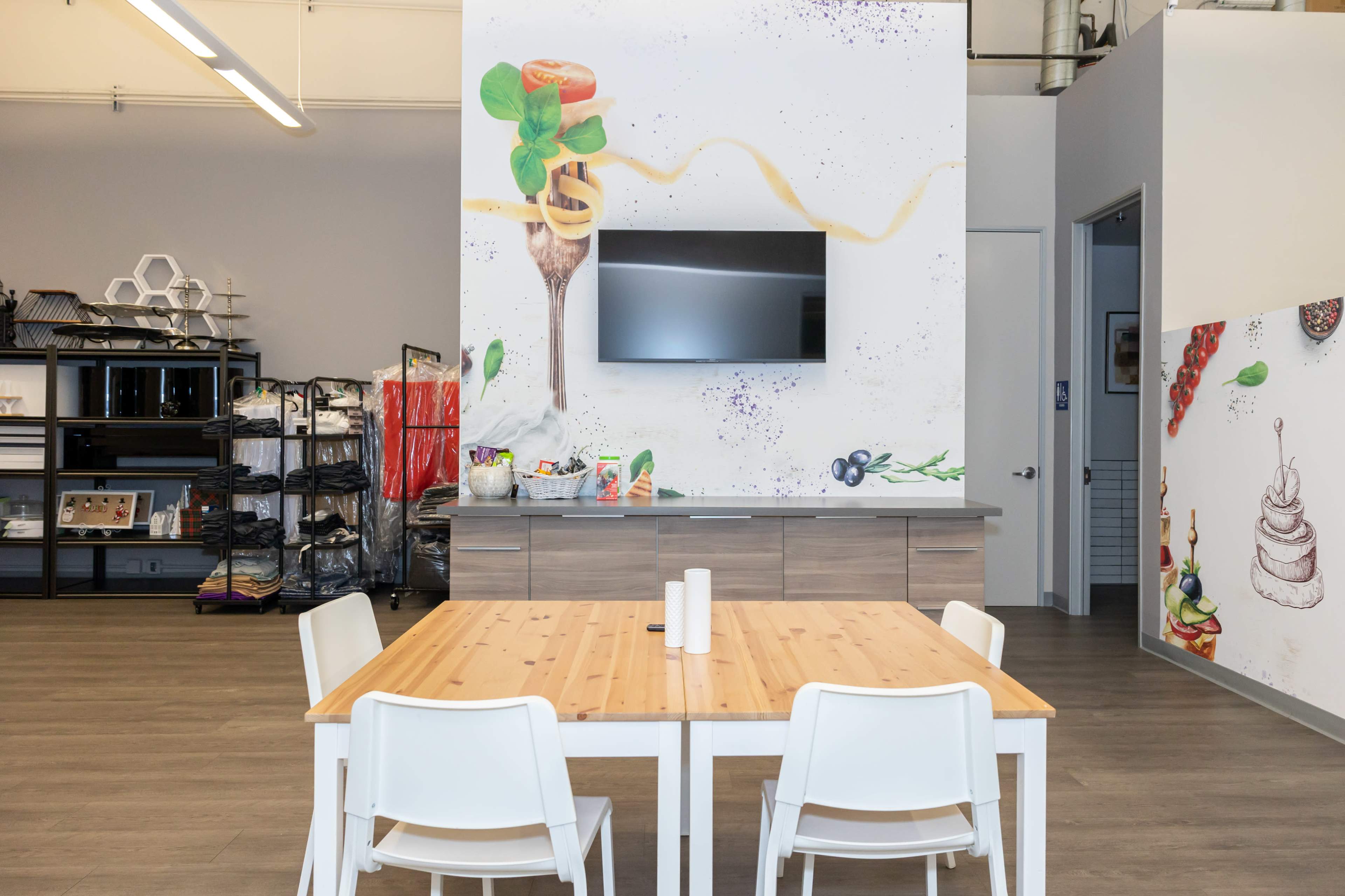 A wooden table with white chairs is set in a room featuring a mural with food-themed illustrations and a mounted television on the wall.