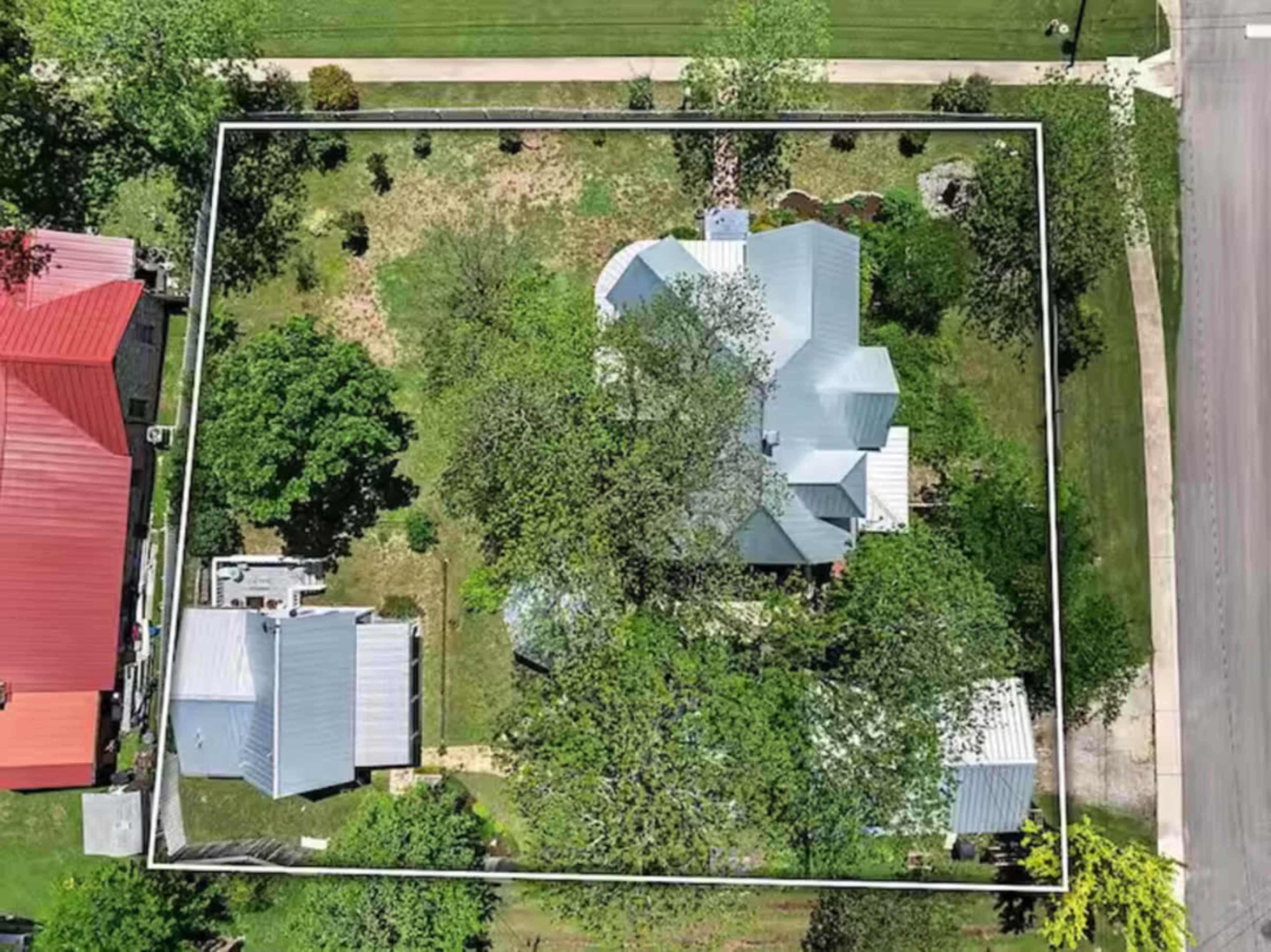 The image shows an aerial view of a residential property surrounded by green trees and grassy areas, with several nearby houses.