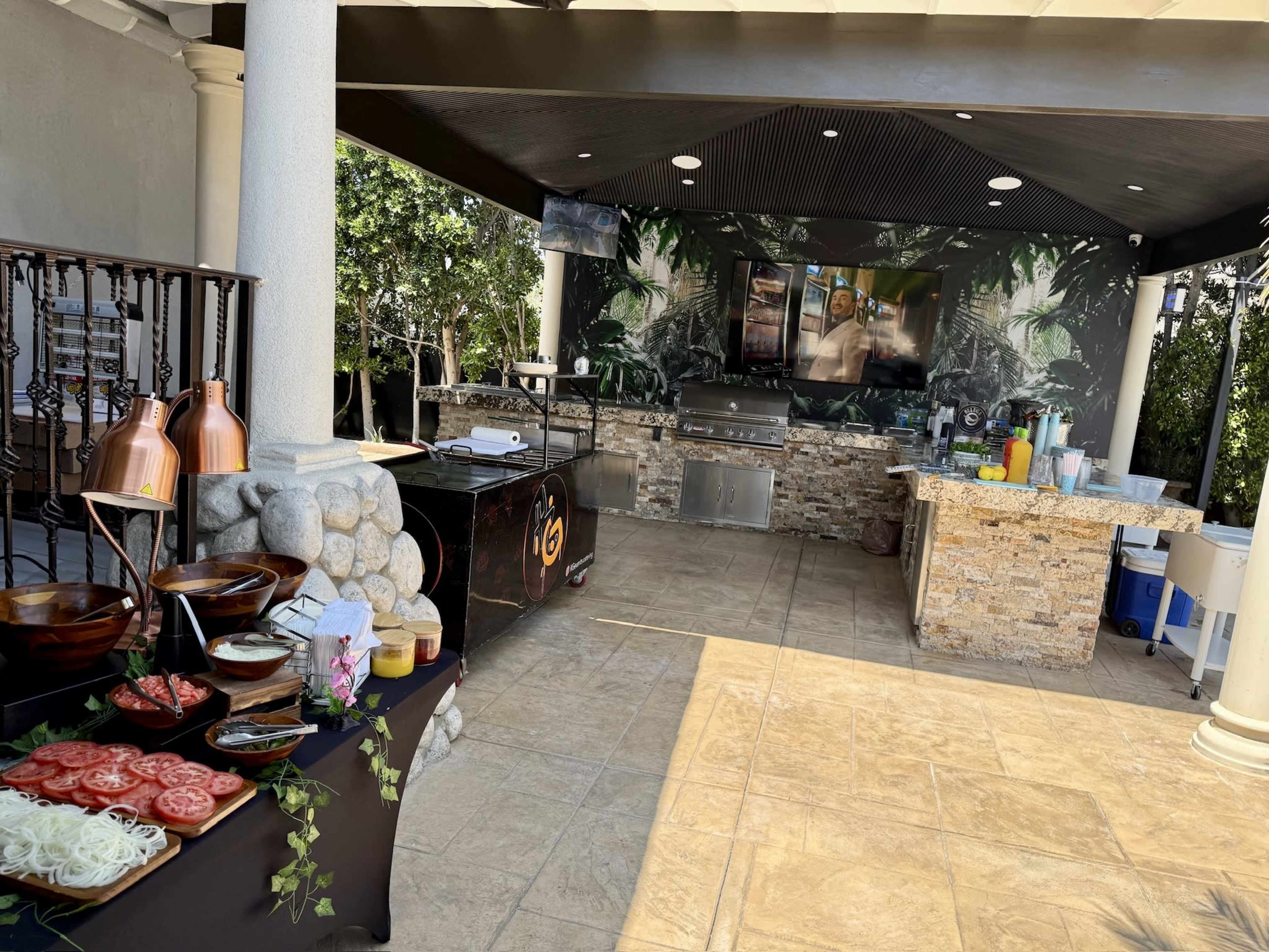The image shows an outdoor kitchen area with a stone grill, a large TV mounted on the wall, and a table displaying fresh ingredients like tomatoes and salads.