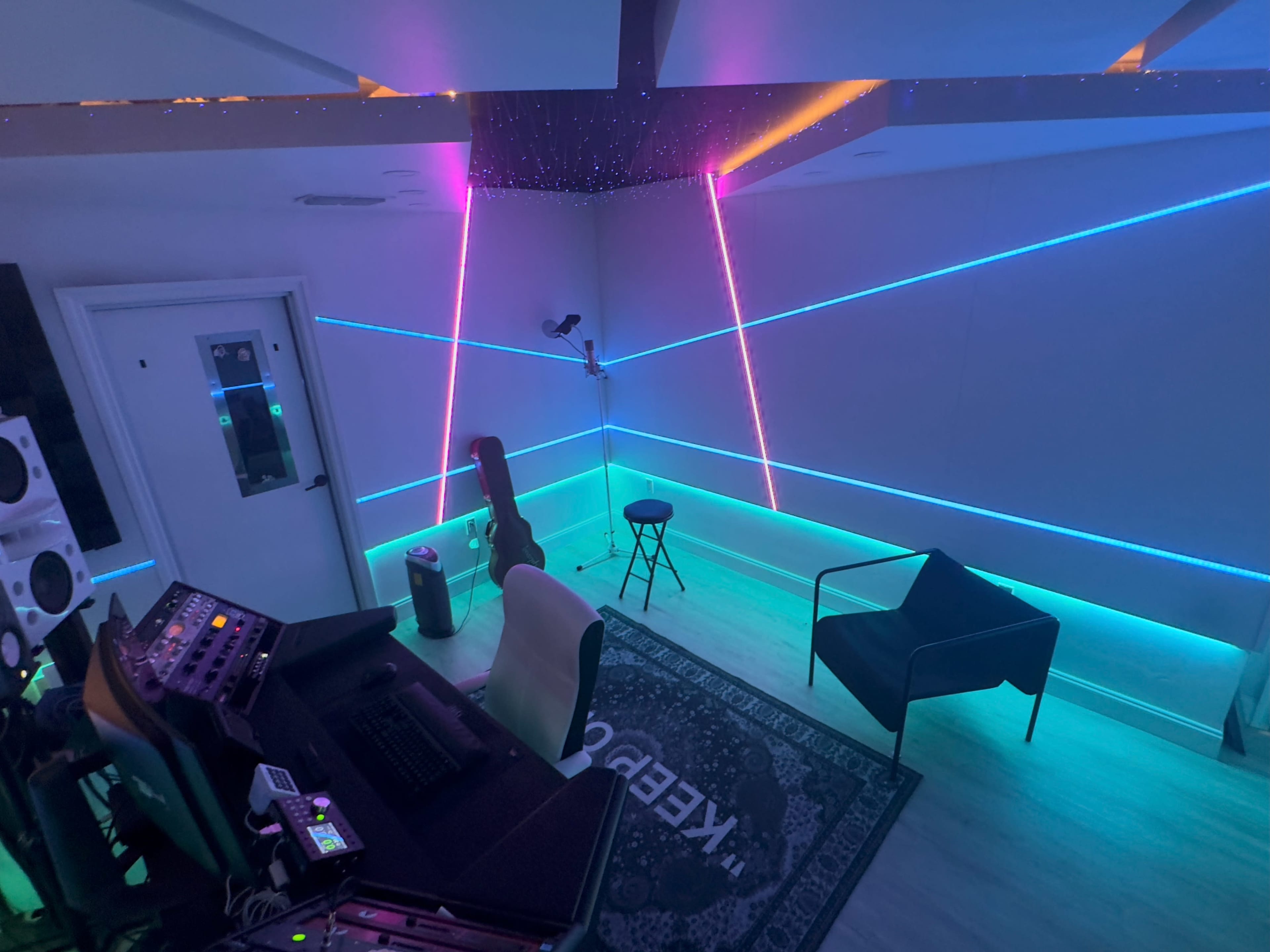 The image shows a dimly lit recording studio featuring colorful LED lighting, a desk with music equipment, and a black chair positioned next to a stool and a guitar.