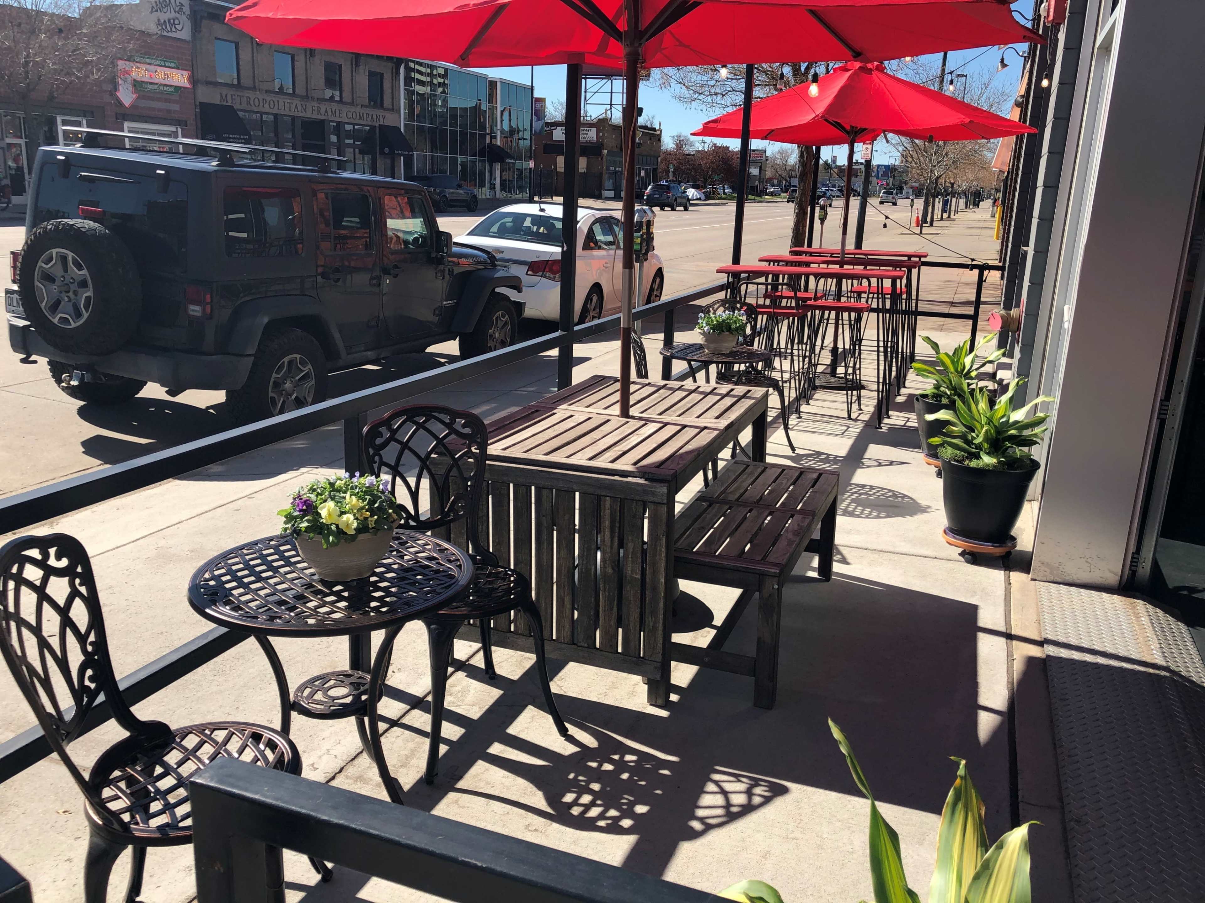 The image shows an outdoor patio with wooden tables and chairs shaded by red umbrellas, alongside a street with parked cars.