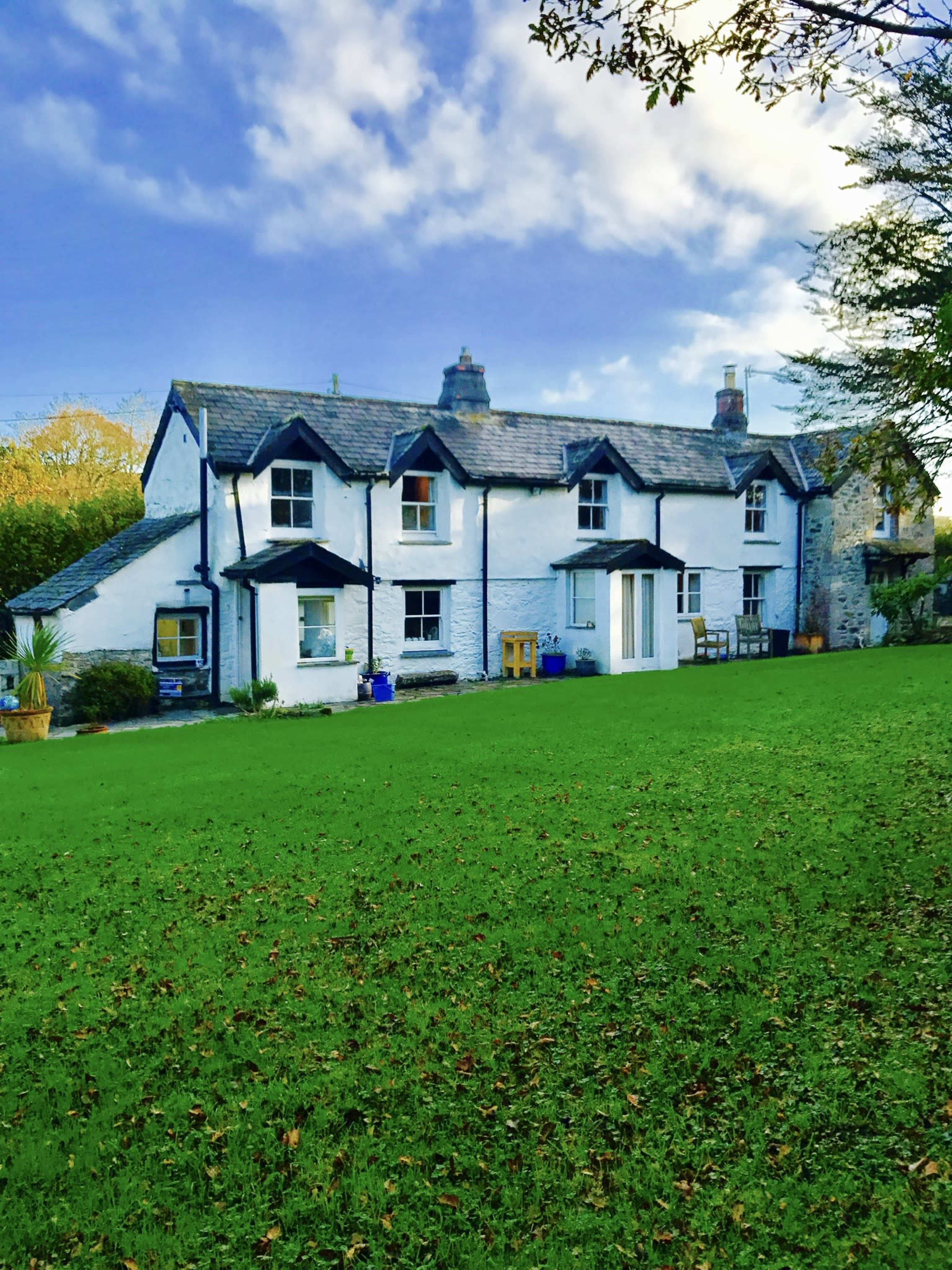 The image shows a two-story white house with a slate roof, surrounded by a green lawn and trees under a partly cloudy sky.