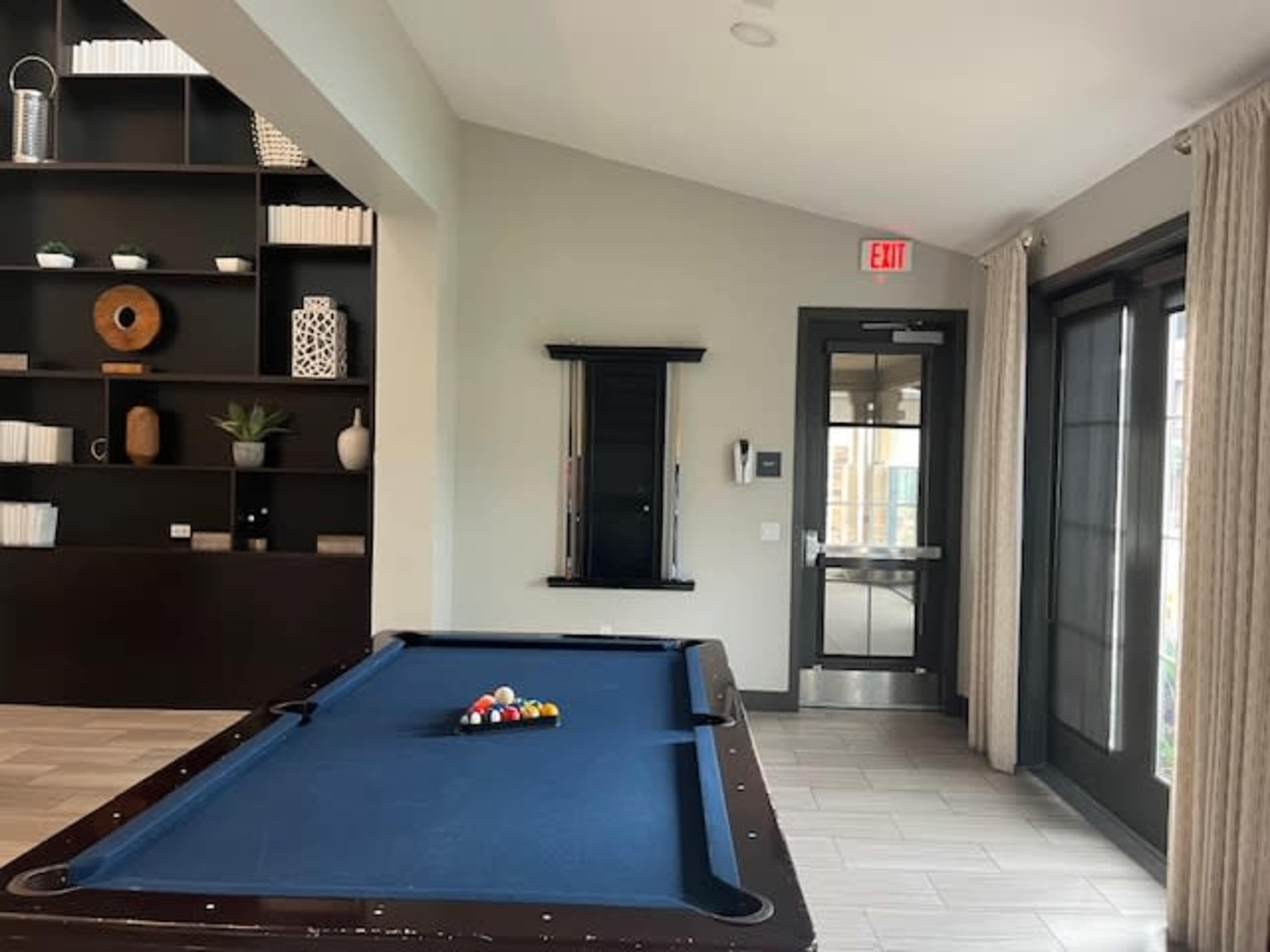 The image shows a billiard table with balls arranged in a triangle, positioned near a wall with a large window and a door leading to an exit.