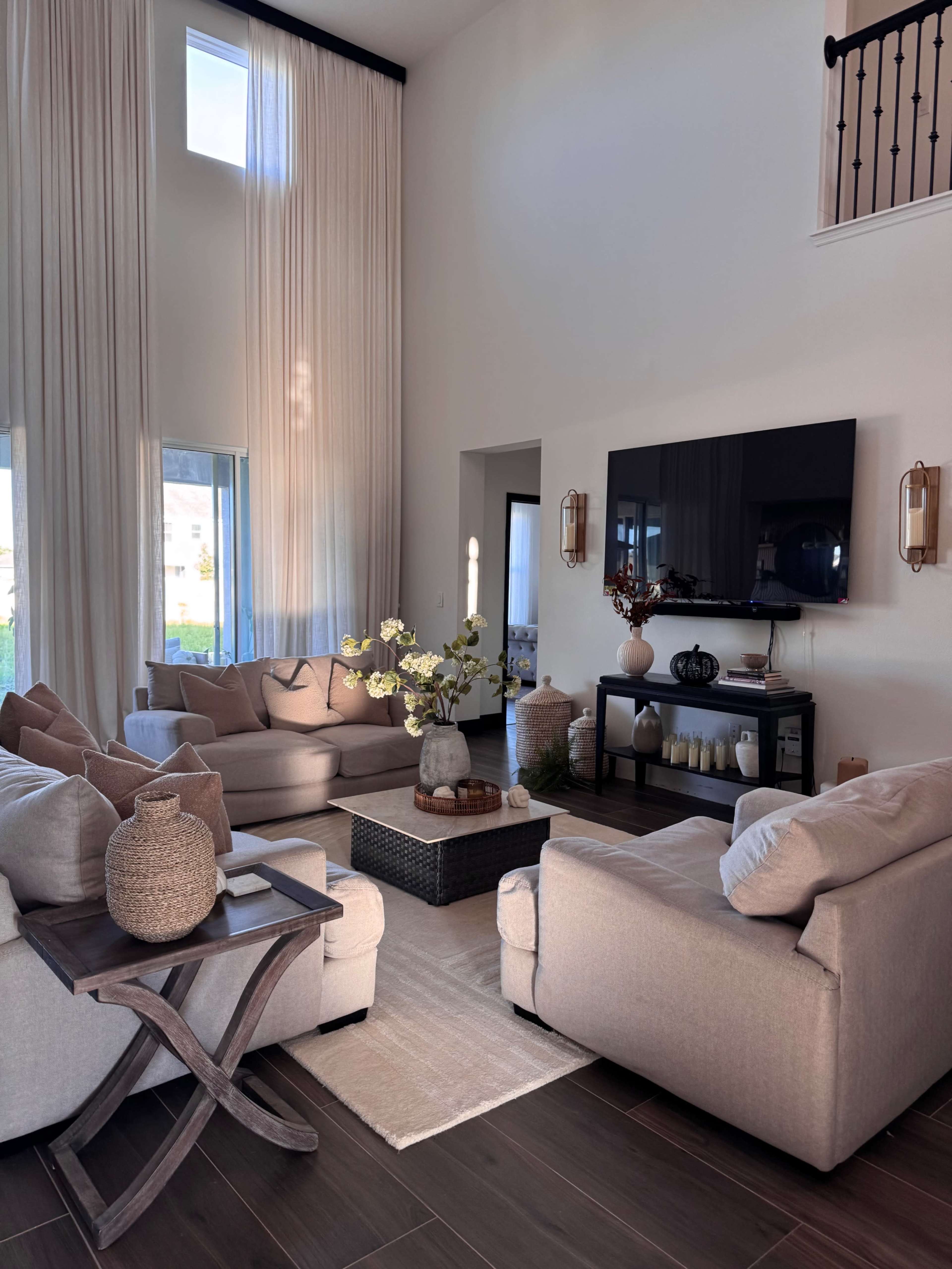 The image shows a spacious living room with large windows, soft seating arranged around a central coffee table, and light-colored decor.