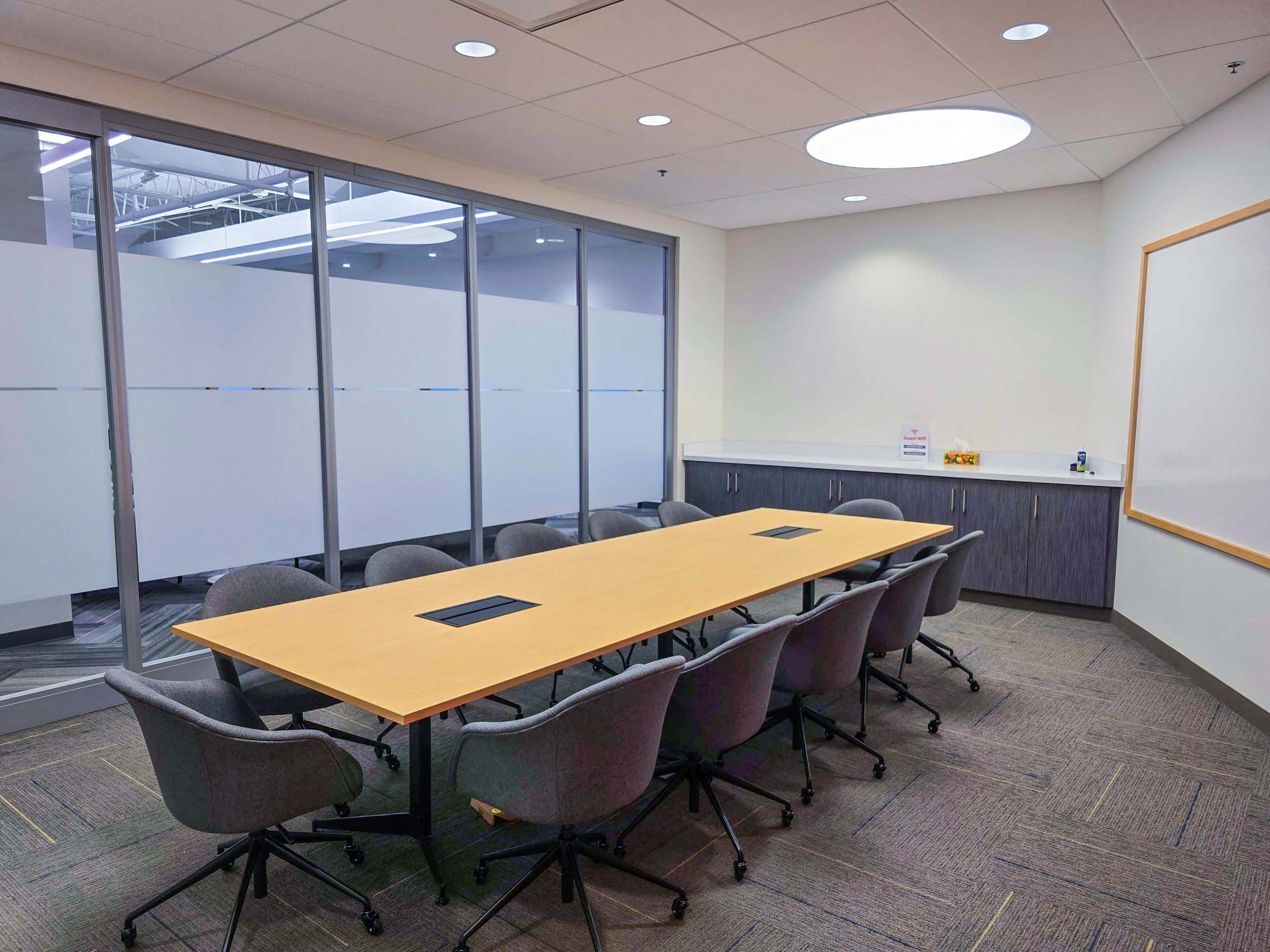 A conference room features a long wooden table surrounded by rolling chairs, with a whiteboard and glass walls that provide visibility into the space.