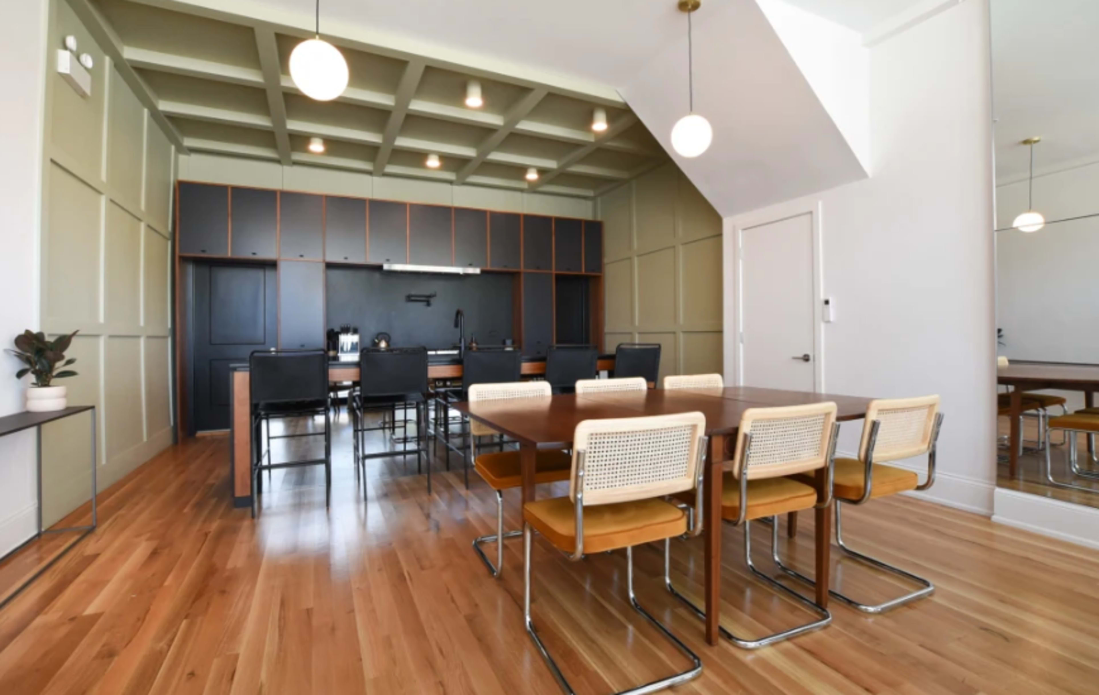 A modern dining room with a long wooden table surrounded by six chairs, and a kitchen area in the background featuring dark cabinets and appliances.