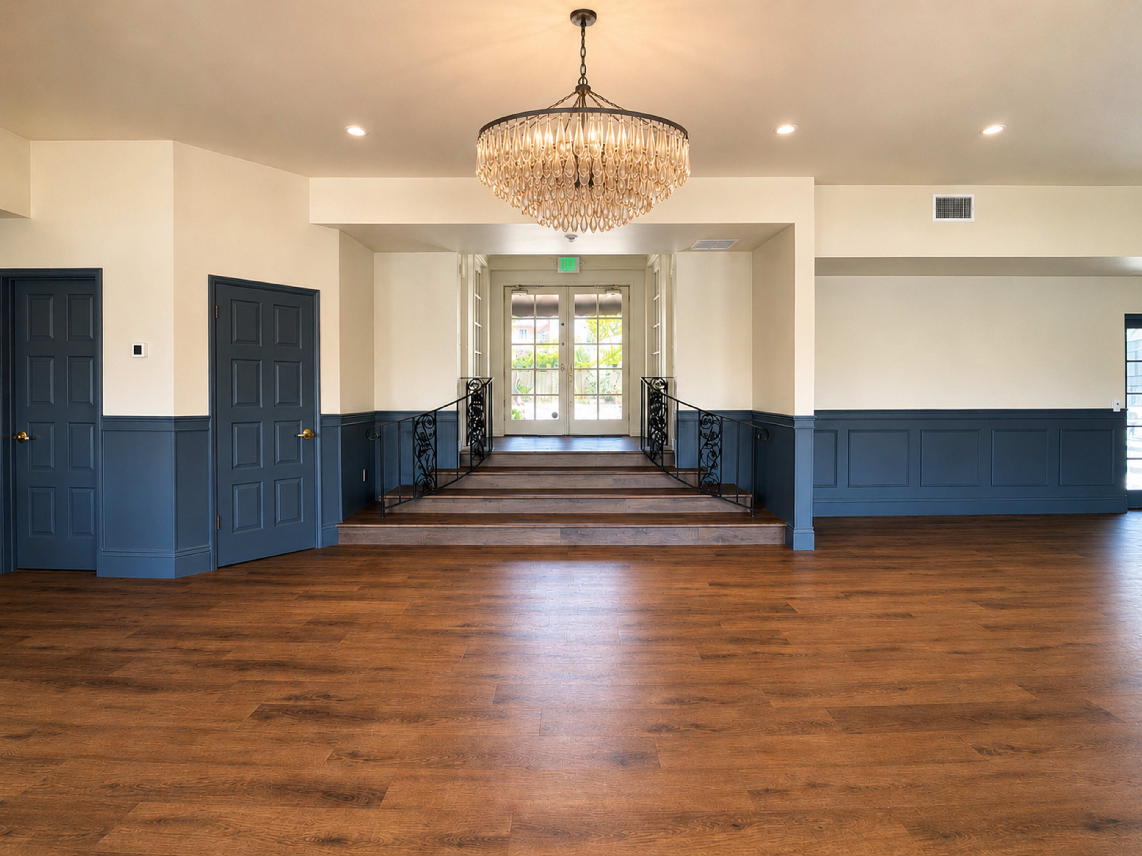 The image shows a spacious interior with a chandelier, wooden flooring, and a staircase leading up to a set of double doors.