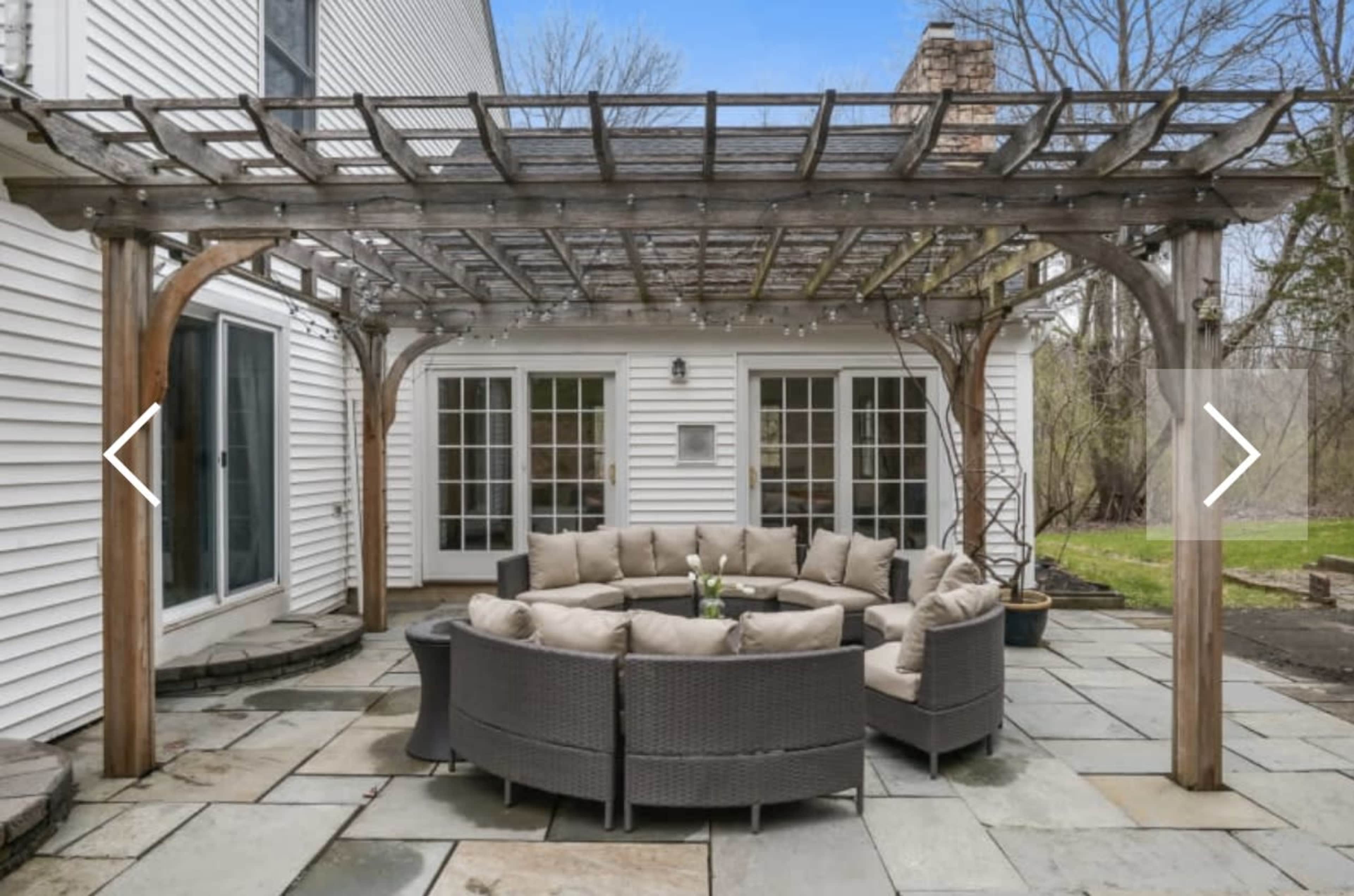 A circular seating area with cushioned chairs is placed under a wooden pergola on a stone patio.
