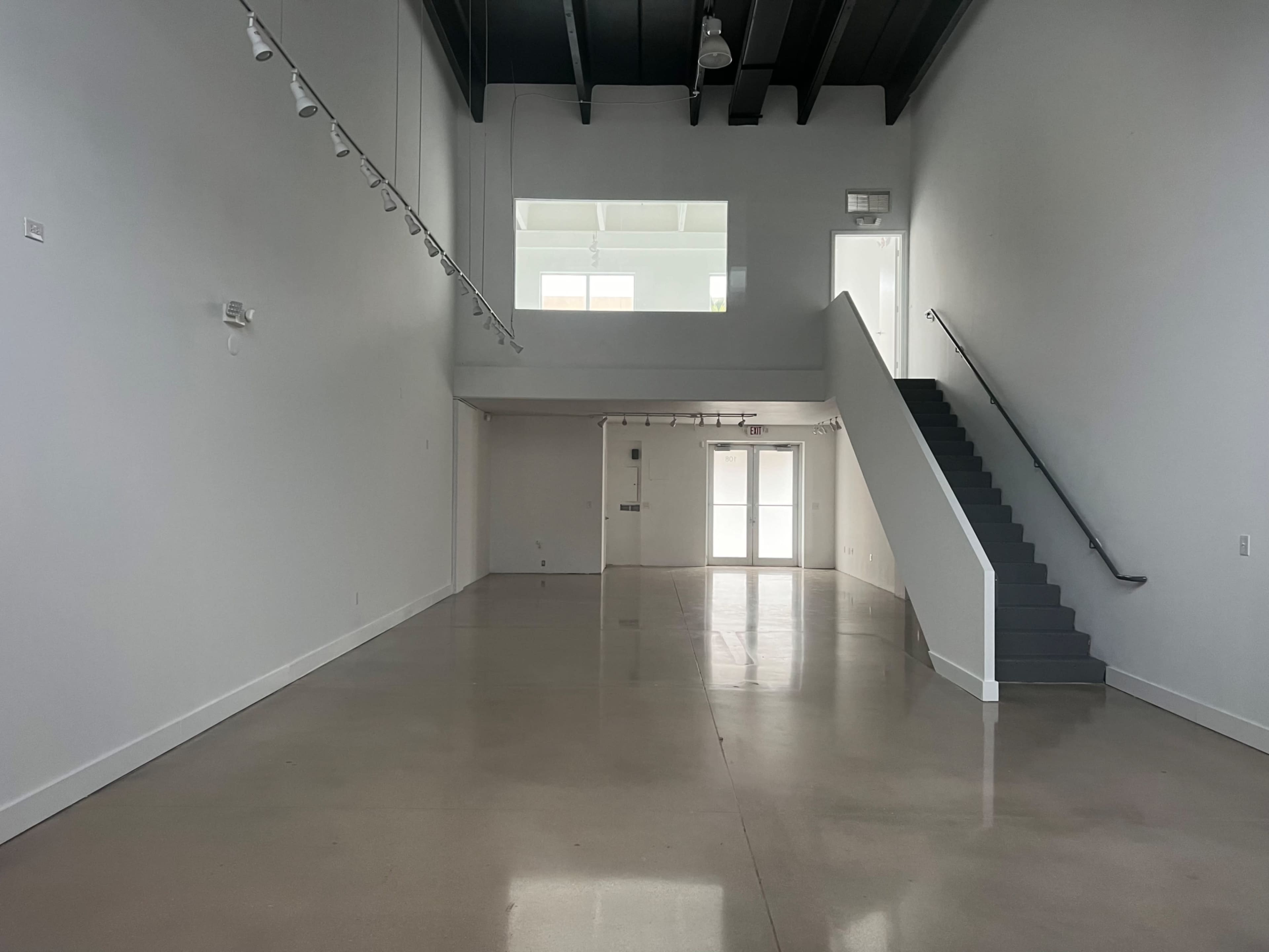 The image shows a spacious, empty interior with polished concrete floors, large windows, and a staircase on one side.