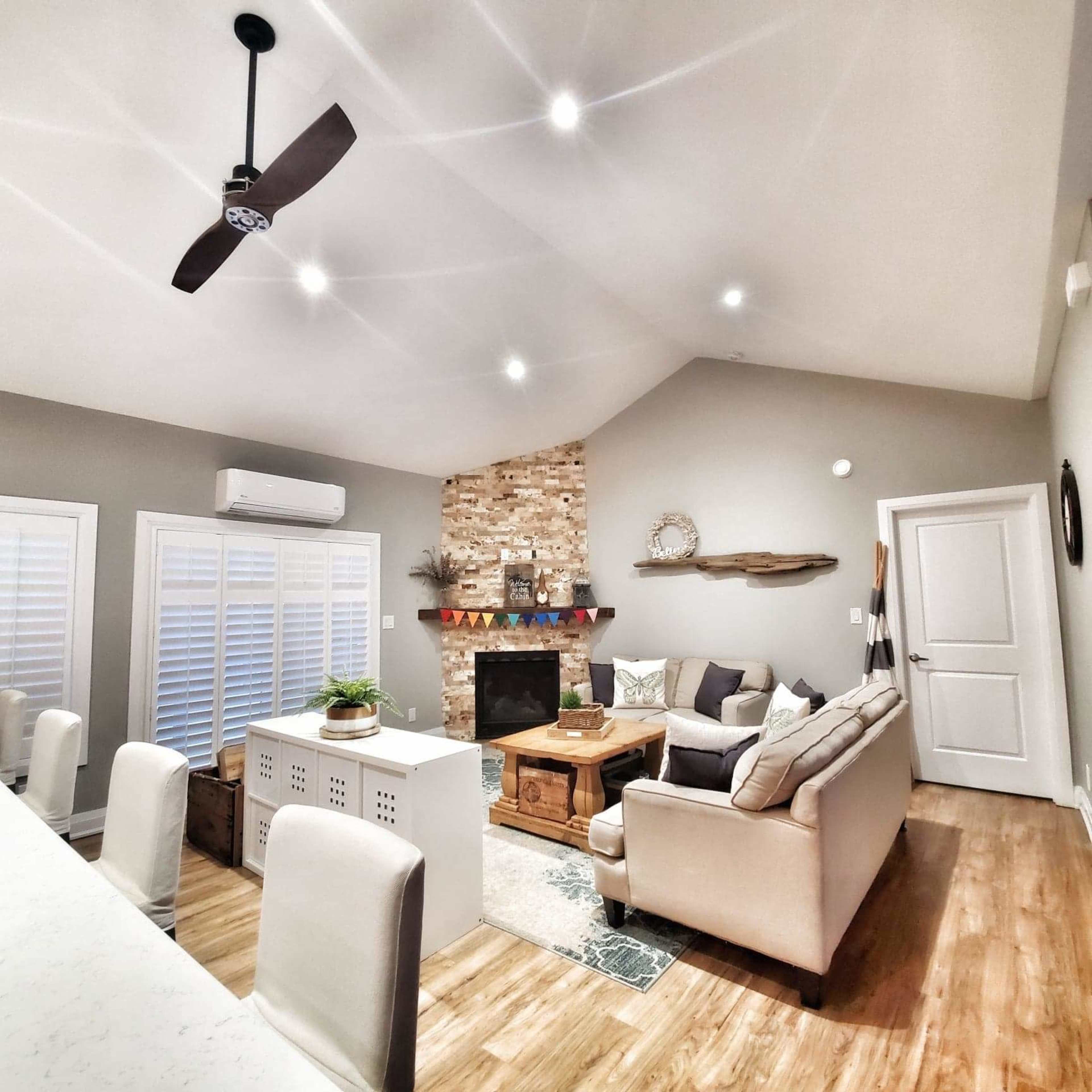 The image shows a modern living room with a stone fireplace, beige sofa, wooden coffee table, and a ceiling fan under a vaulted ceiling.