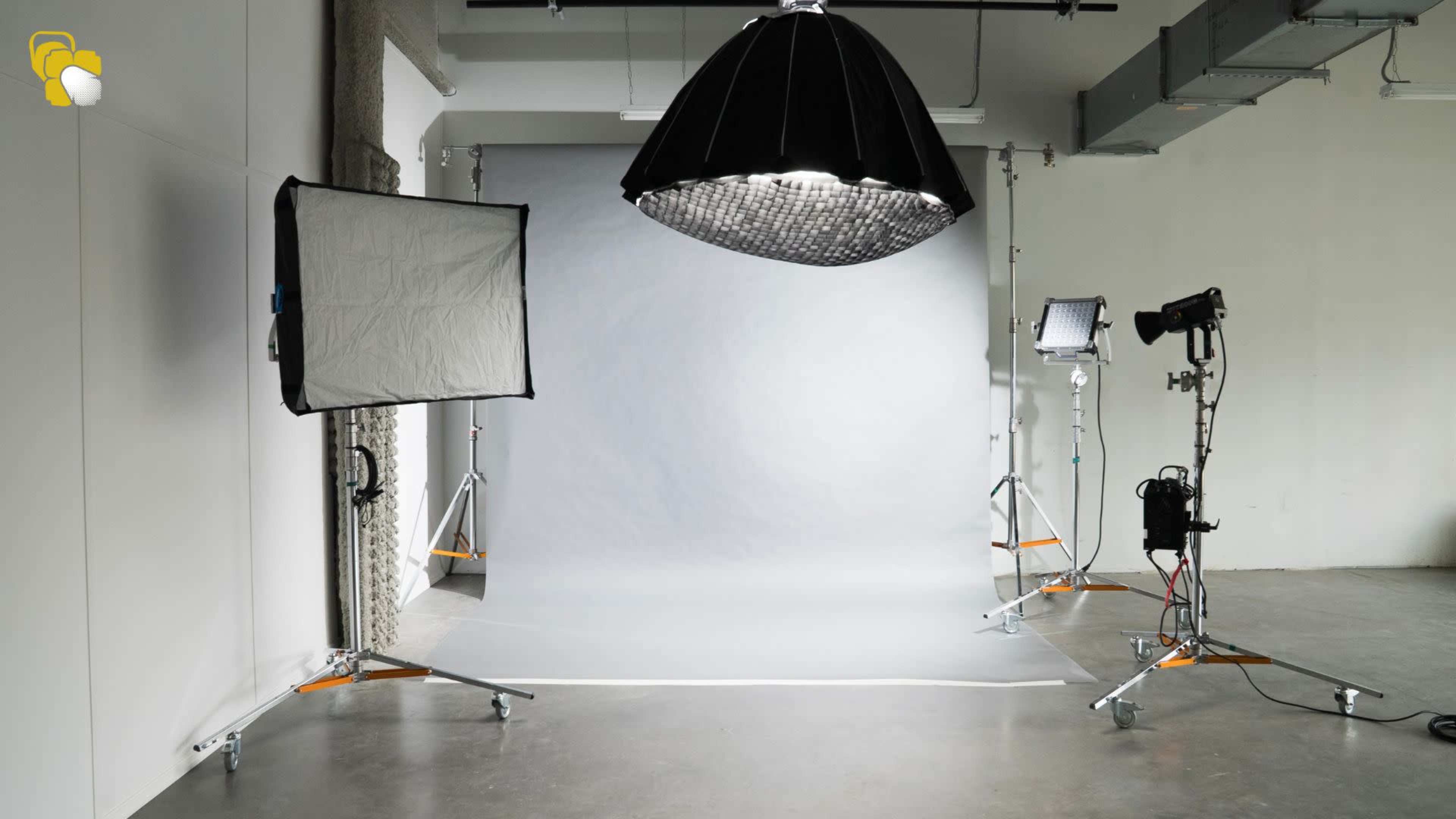 The image shows a photography studio setup featuring a gray backdrop, softbox lights, and a large hanging umbrella light.