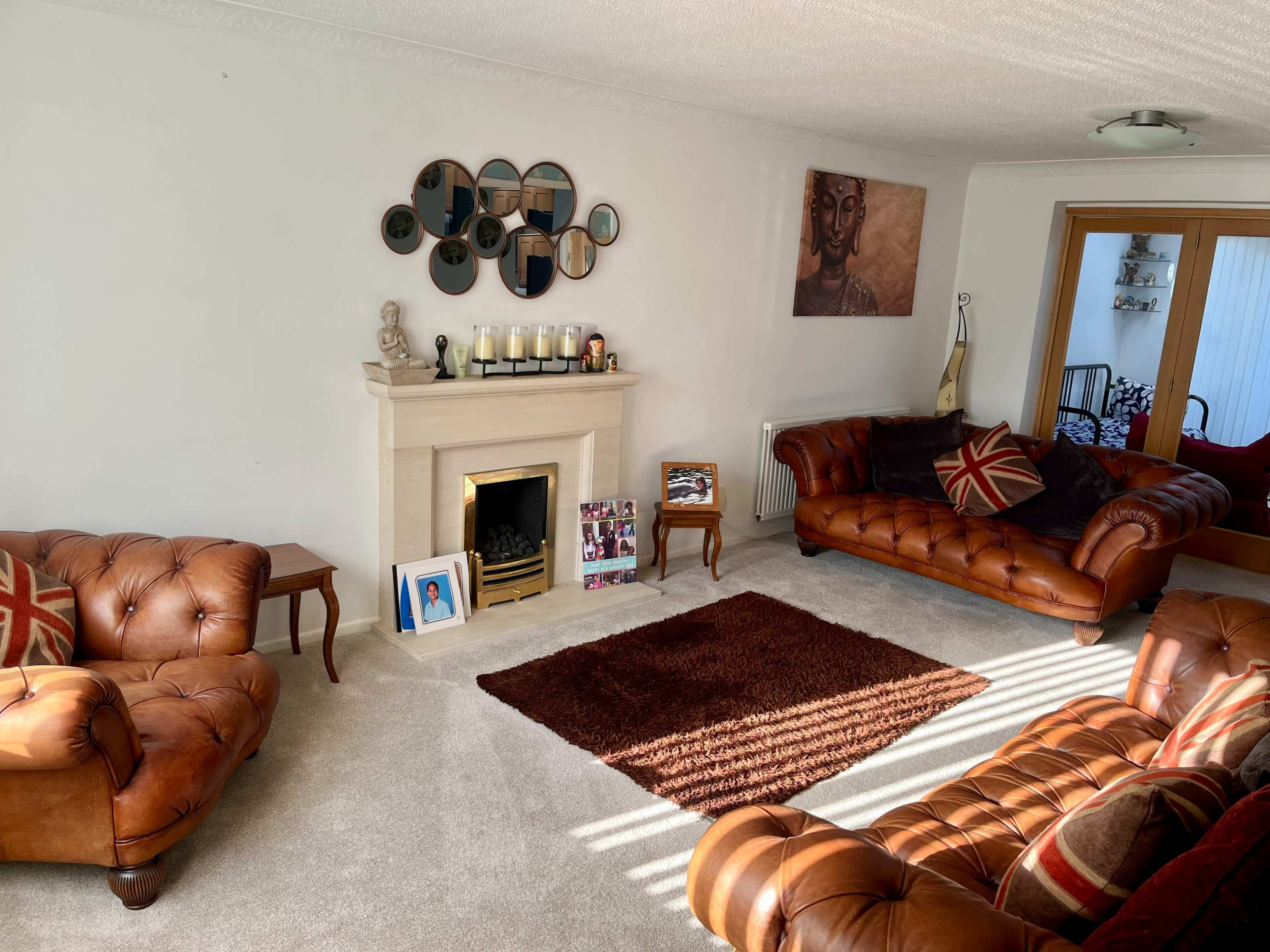 The living room features two brown leather sofas, a fireplace with candles, and a decorative wall mirror arrangement opposite a carpeted area.