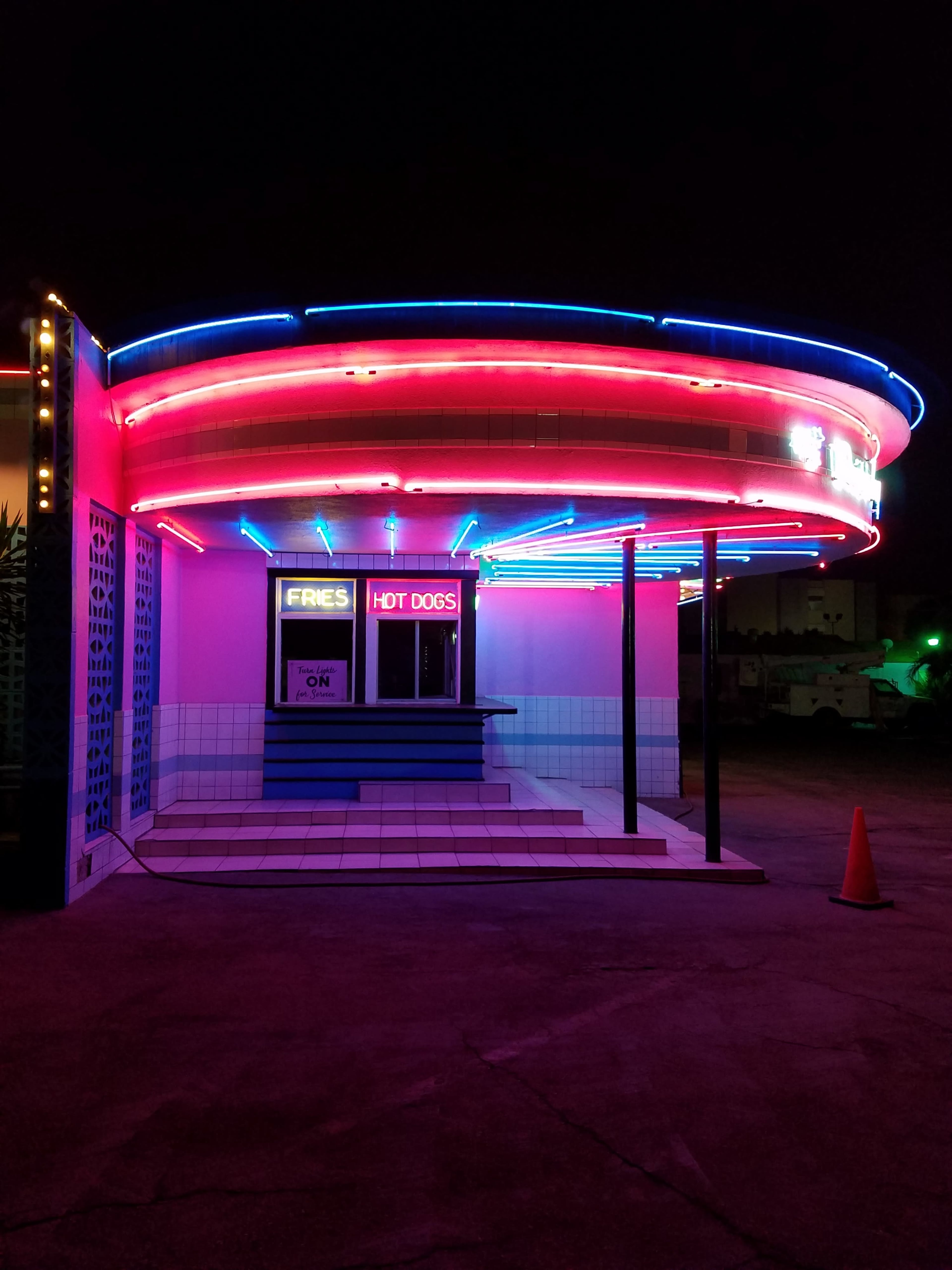 A brightly lit hot dog stand with neon blue and pink lights is centered in the image against a dark night sky.