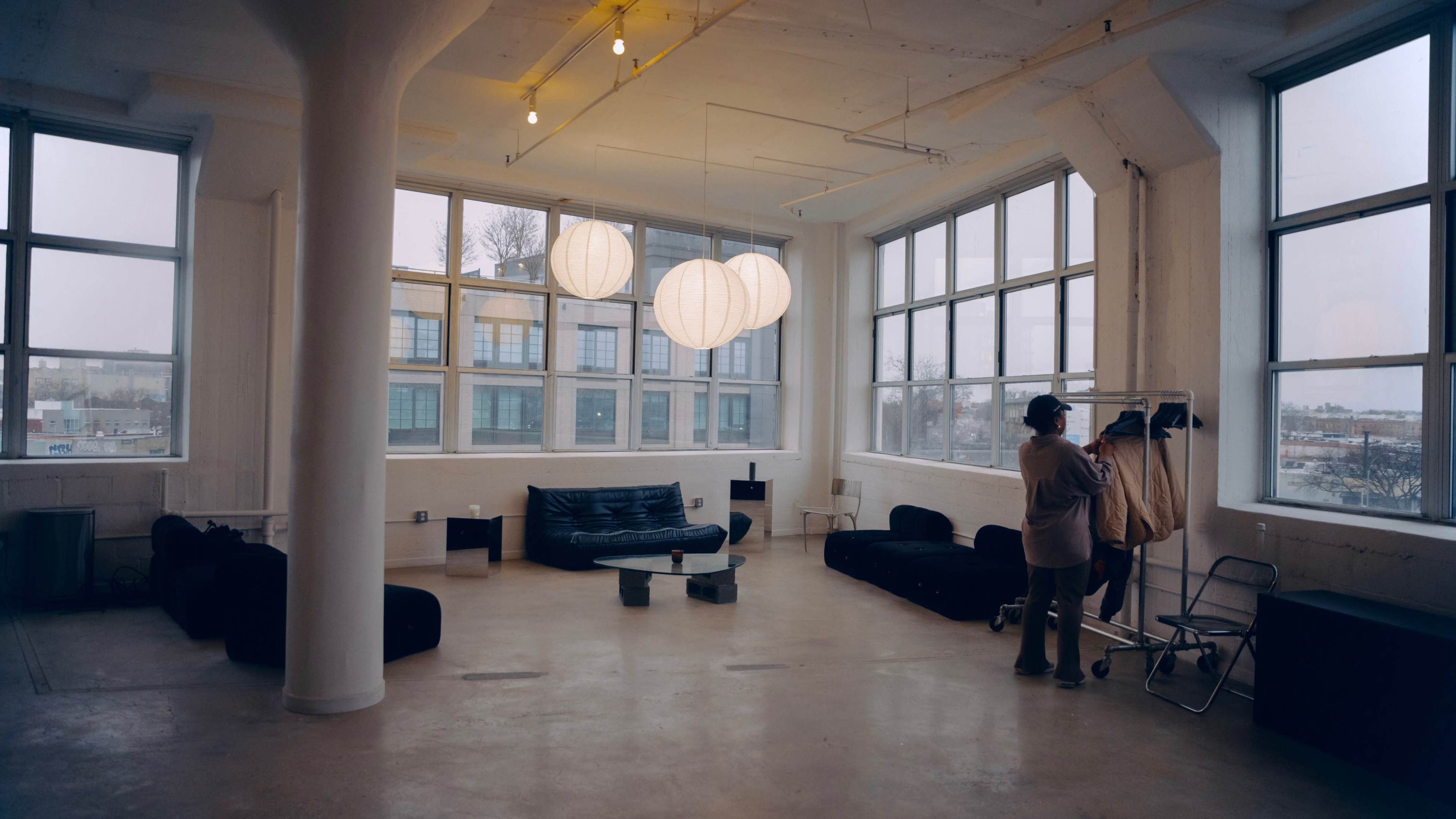 A person stands beside a clothing rack in a spacious, industrial-style room with large windows and modern furniture.
