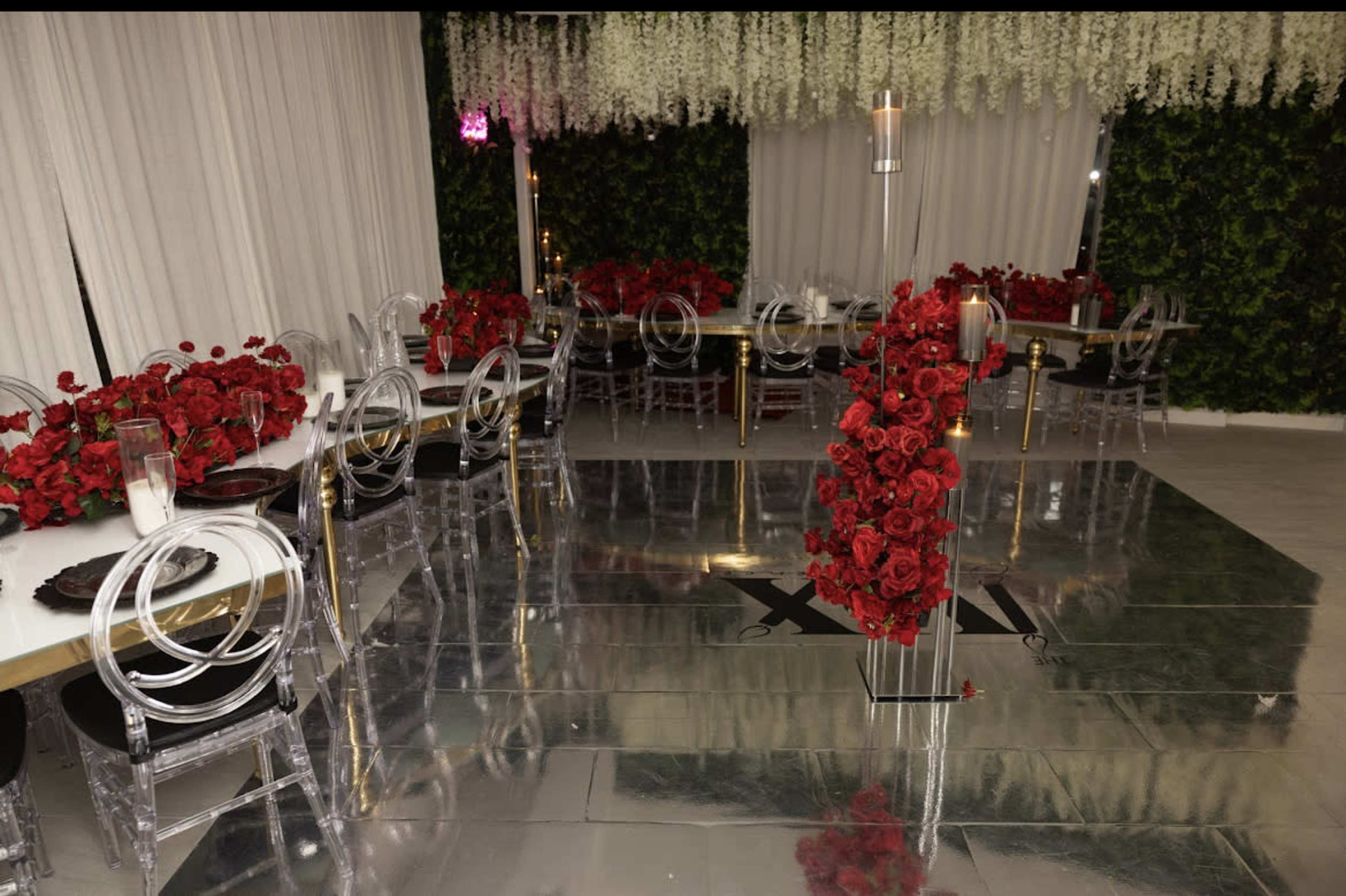 A dining area features clear acrylic chairs and tables adorned with red rose centerpieces and floral arrangements, against a backdrop of white curtains and green foliage.