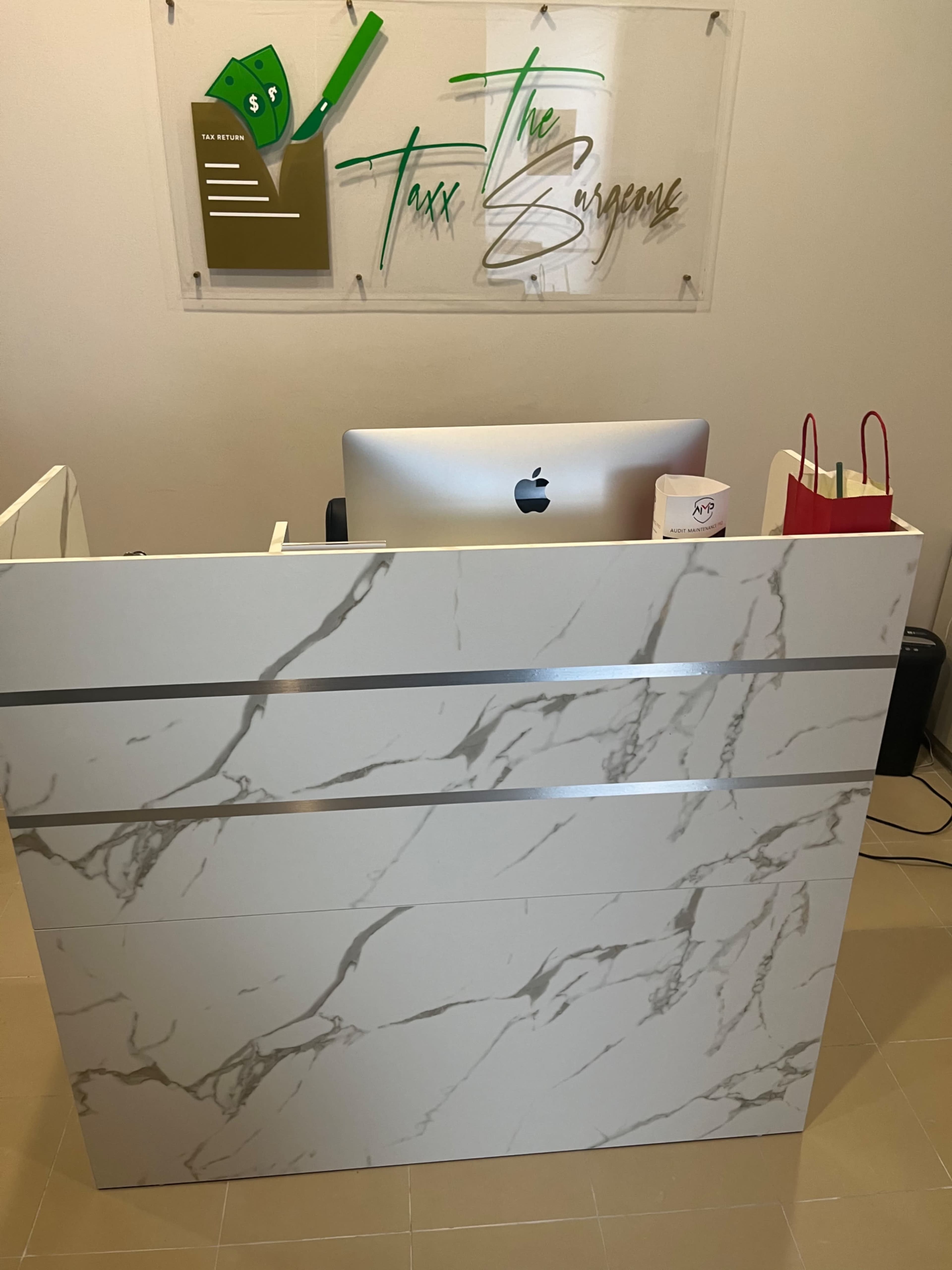 The image shows a modern reception desk with a marble design, positioned in front of a sign that reads "Taxx The Surgeons."