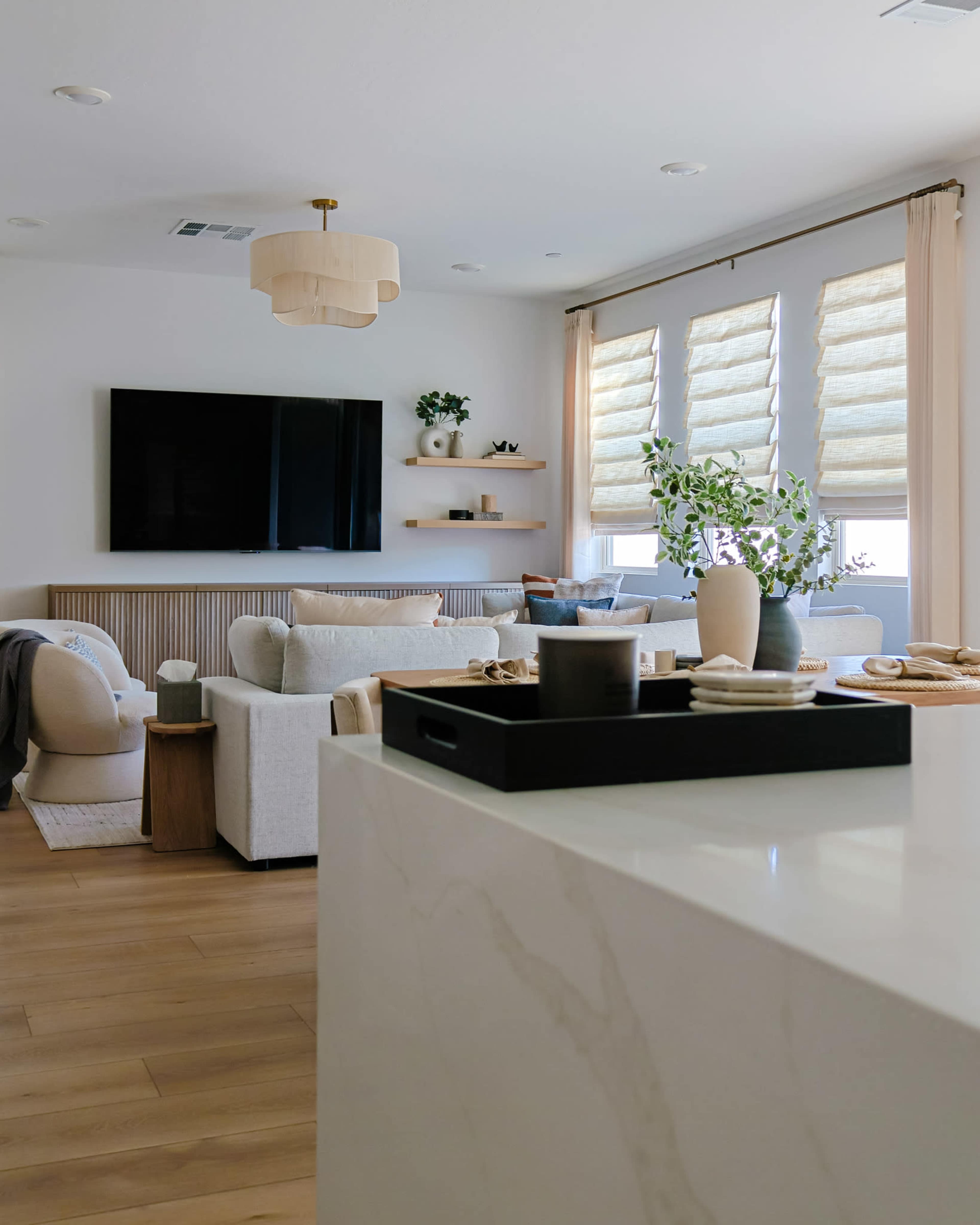 The image depicts a modern living room with a large television, light-colored furniture, and decorative elements arranged on shelves.