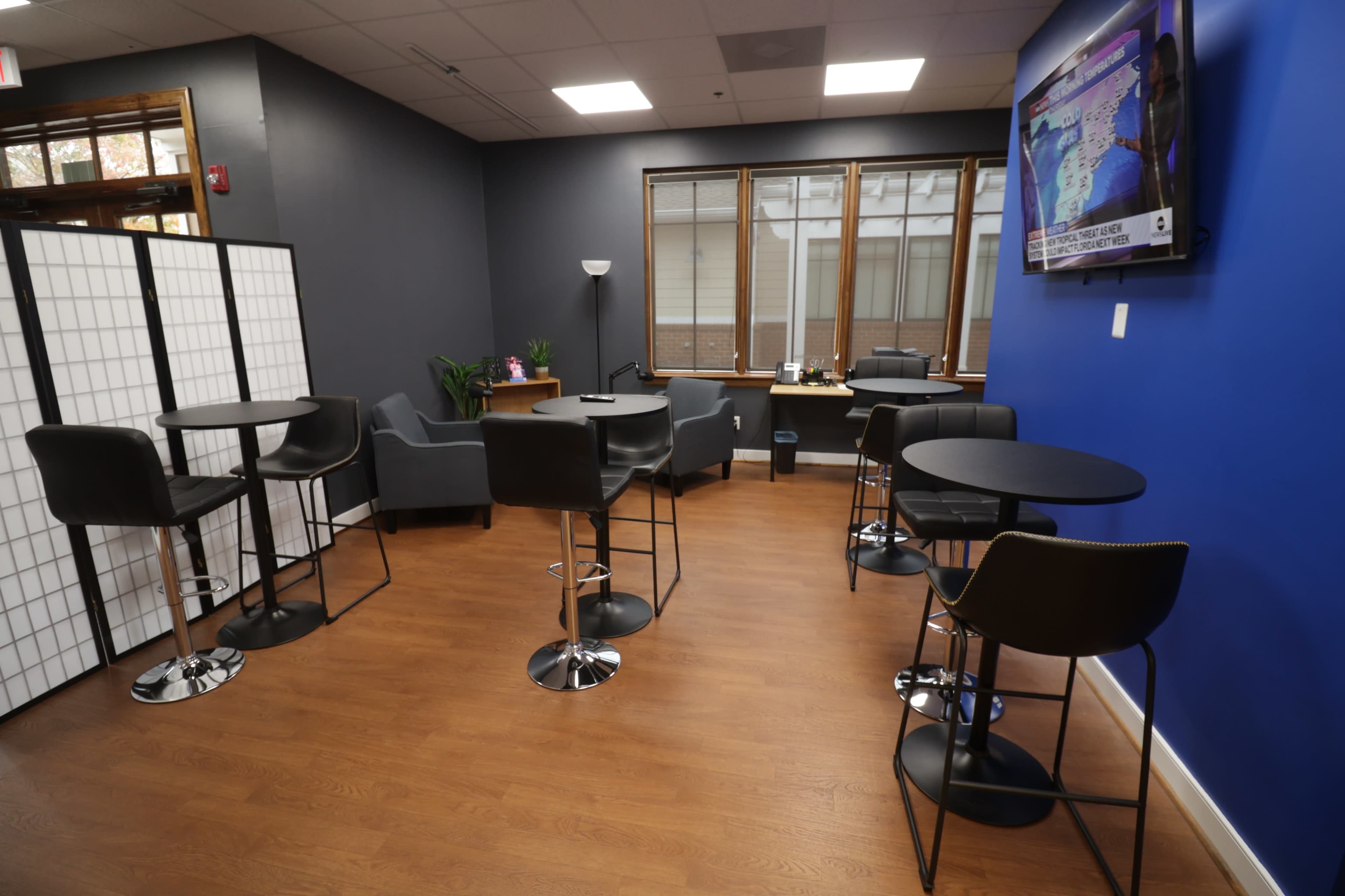 The image shows a modern lounge area with gray seating, tall black stools, and small round tables against blue and white walls, featuring a television on one side.