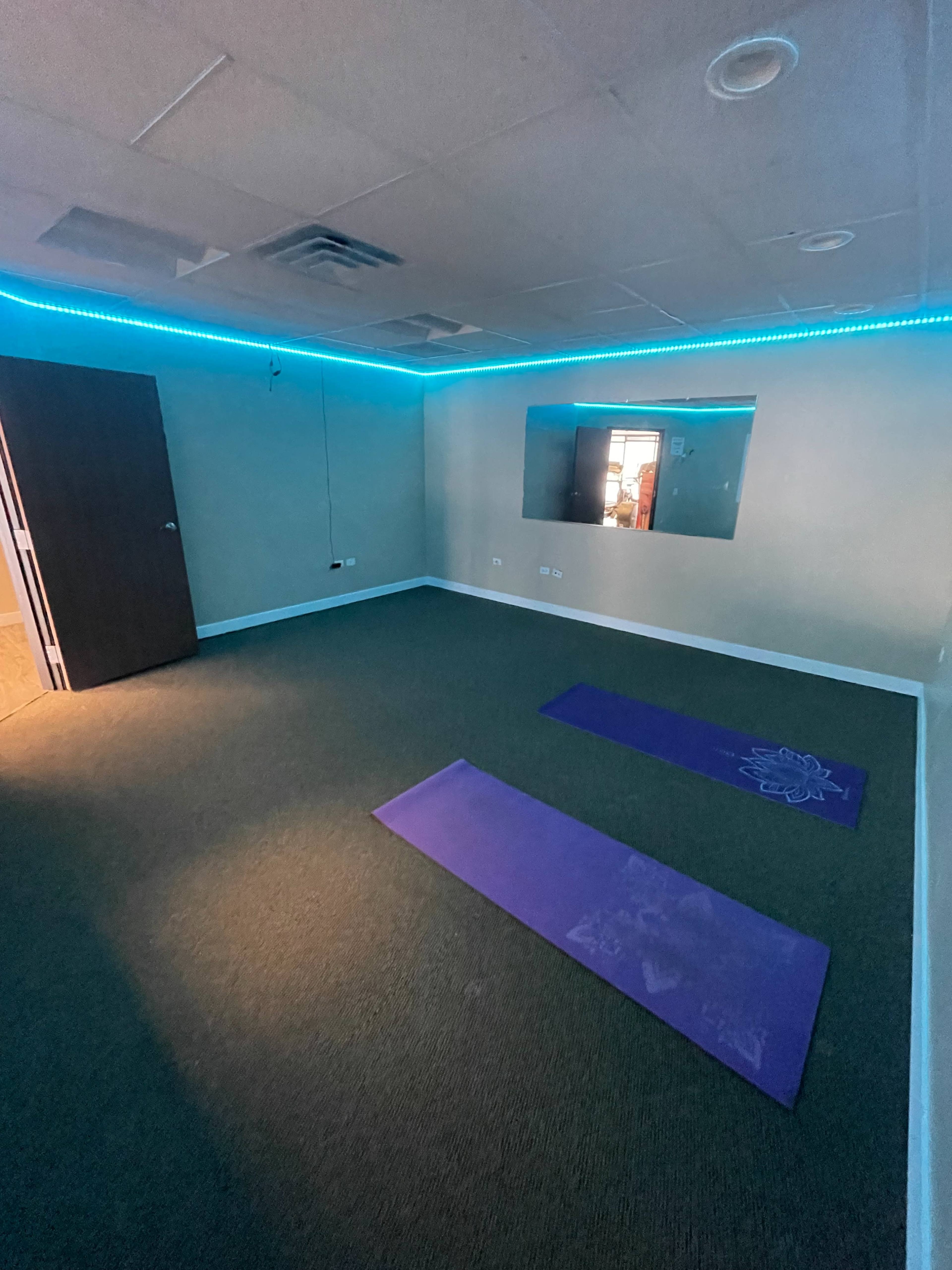 A spacious yoga room with purple mats on a carpeted floor, illuminated by blue ambient lighting along the ceiling.