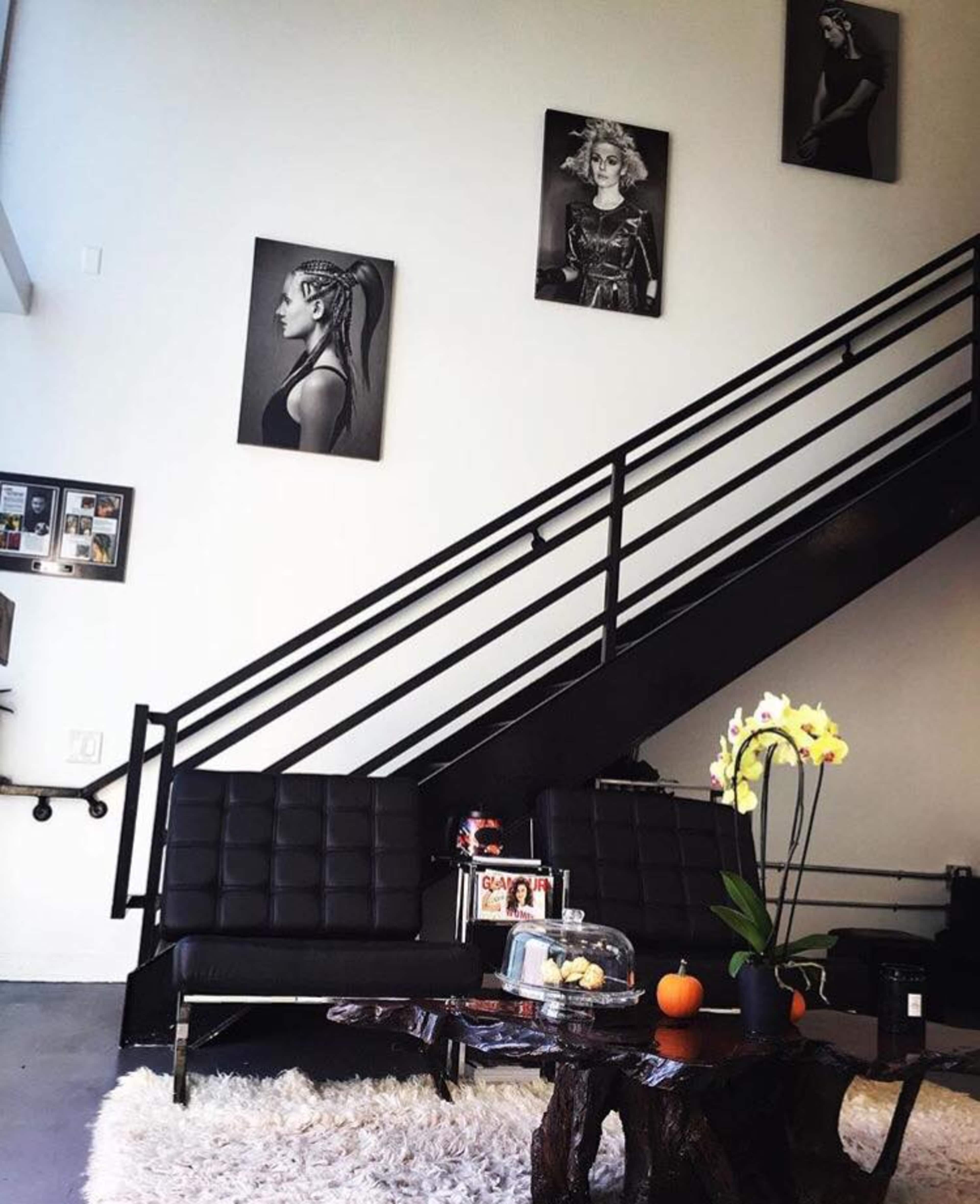 A modern interior space with black seating, a decorative wooden table, an orange pumpkin, and framed portraits on the wall, along with a staircase in the background.