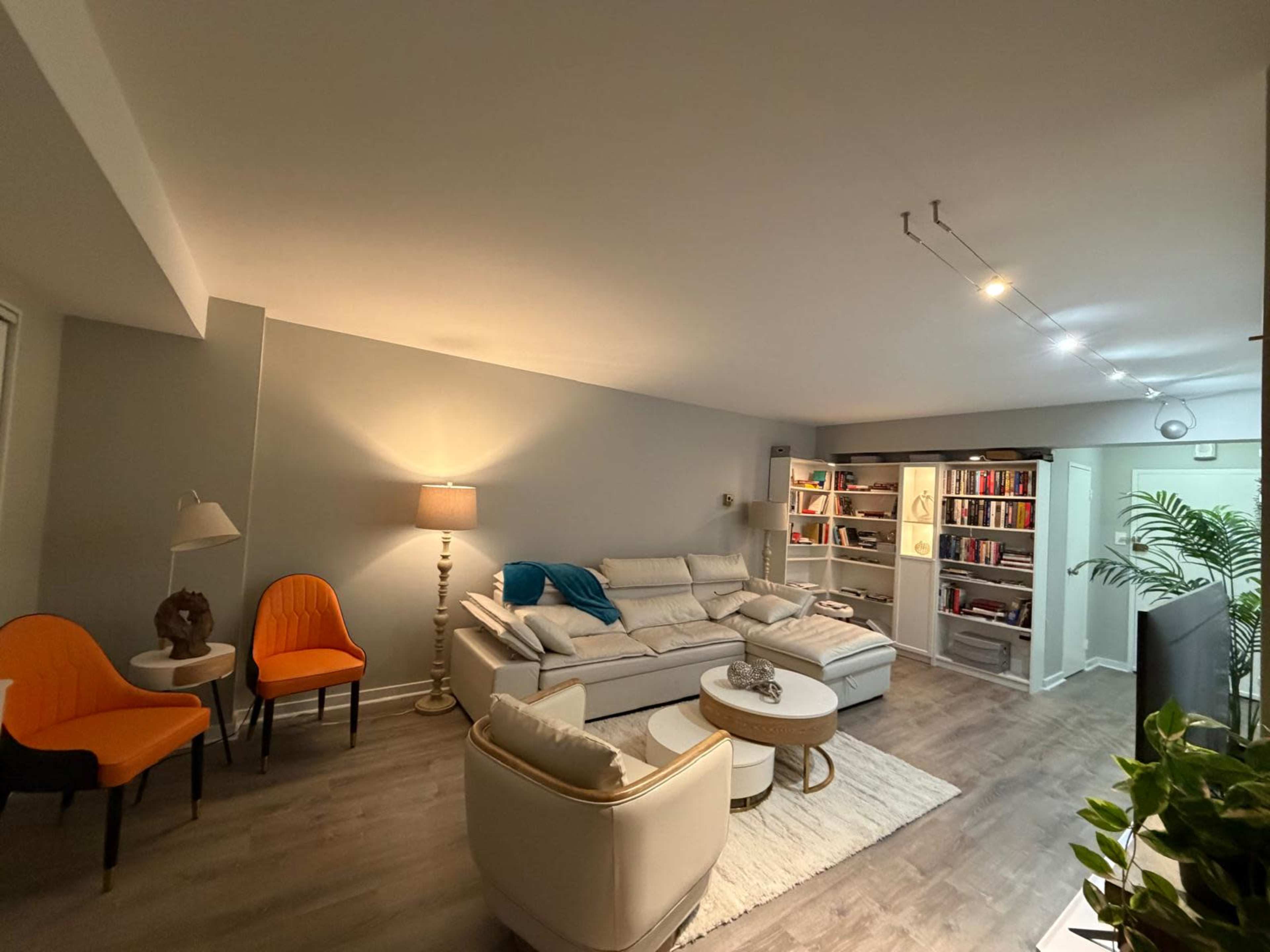 The image shows a modern living room with a sectional sofa, a lamp, two orange chairs, a bookshelf filled with books, and a potted plant.