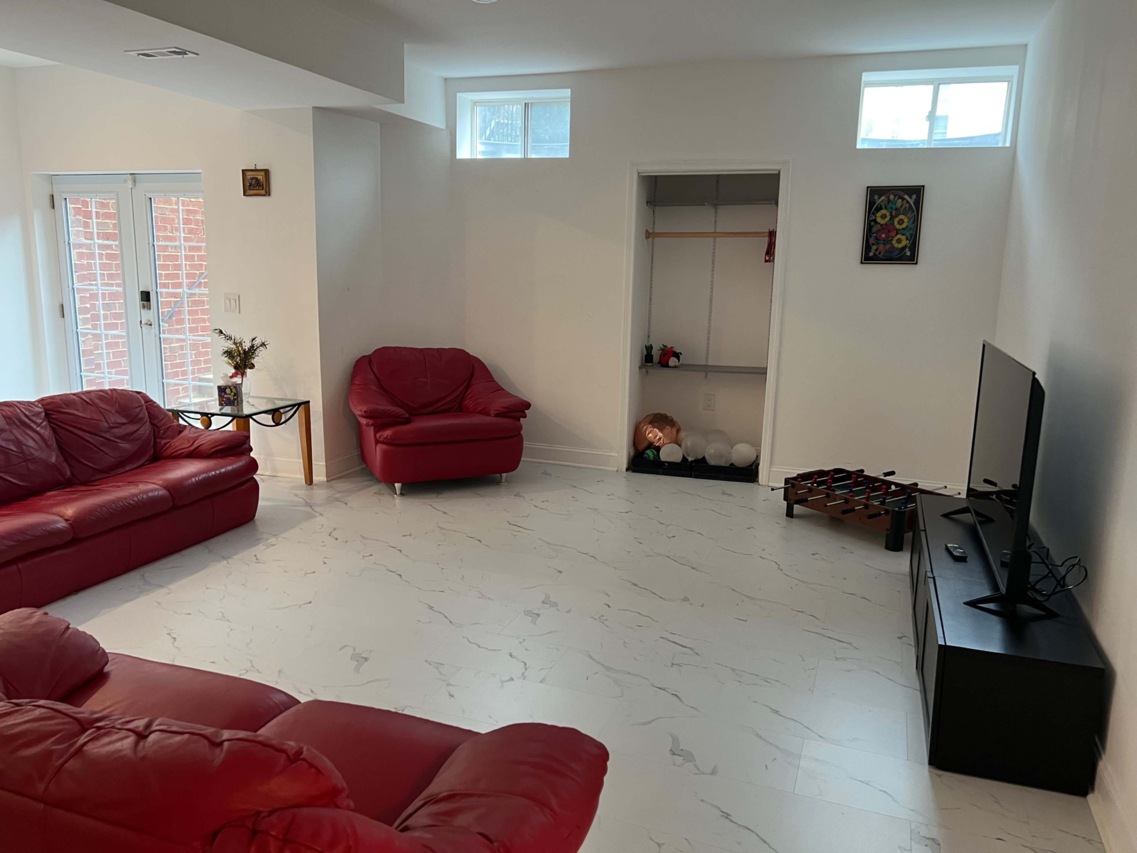 The room features red leather couches, a television on a black stand, and a small closet with storage, all set against a backdrop of white walls and a marble-patterned floor.