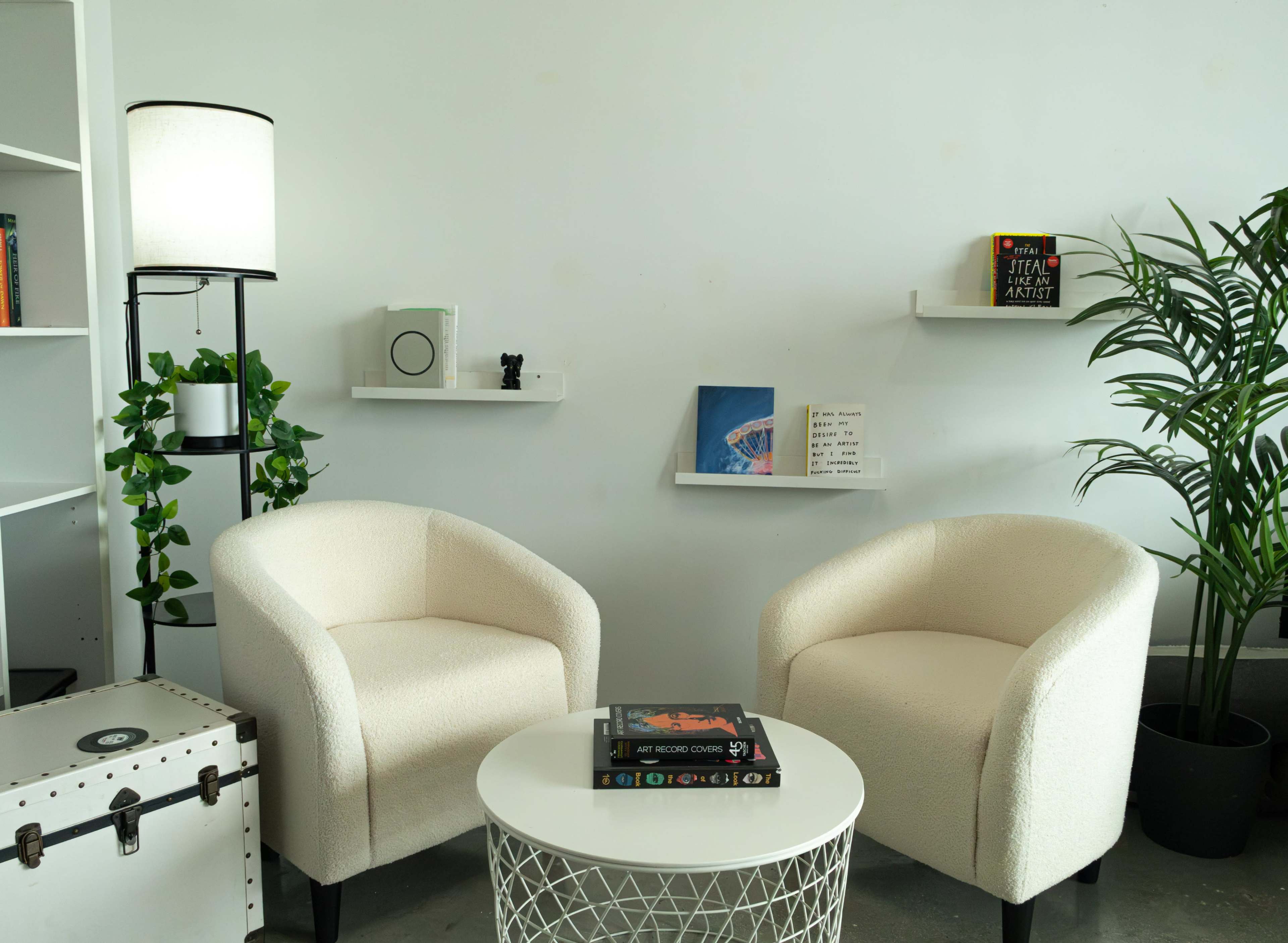 The image shows a minimalist seating area featuring two white upholstered chairs, a round table, a potted plant, and wall shelves with decorative items against a neutral wall.