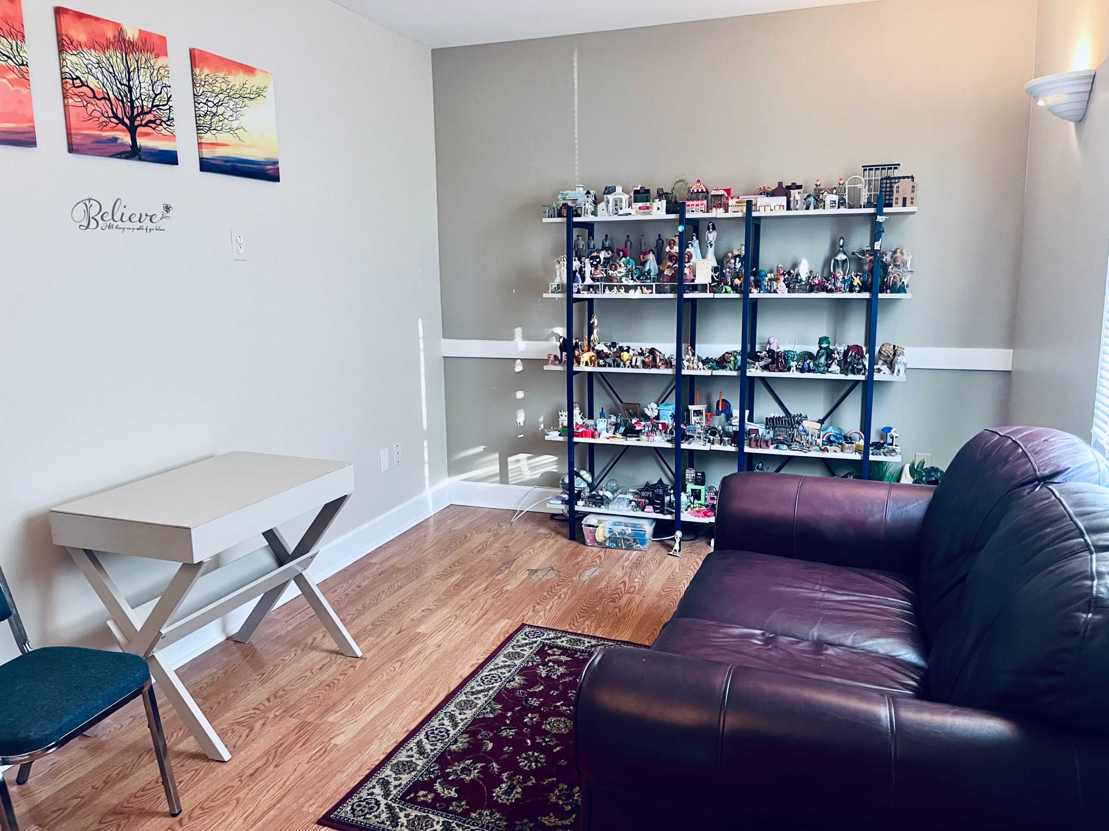 A living room features a brown couch, a small white table, and a shelf filled with decorative collectibles along the wall.