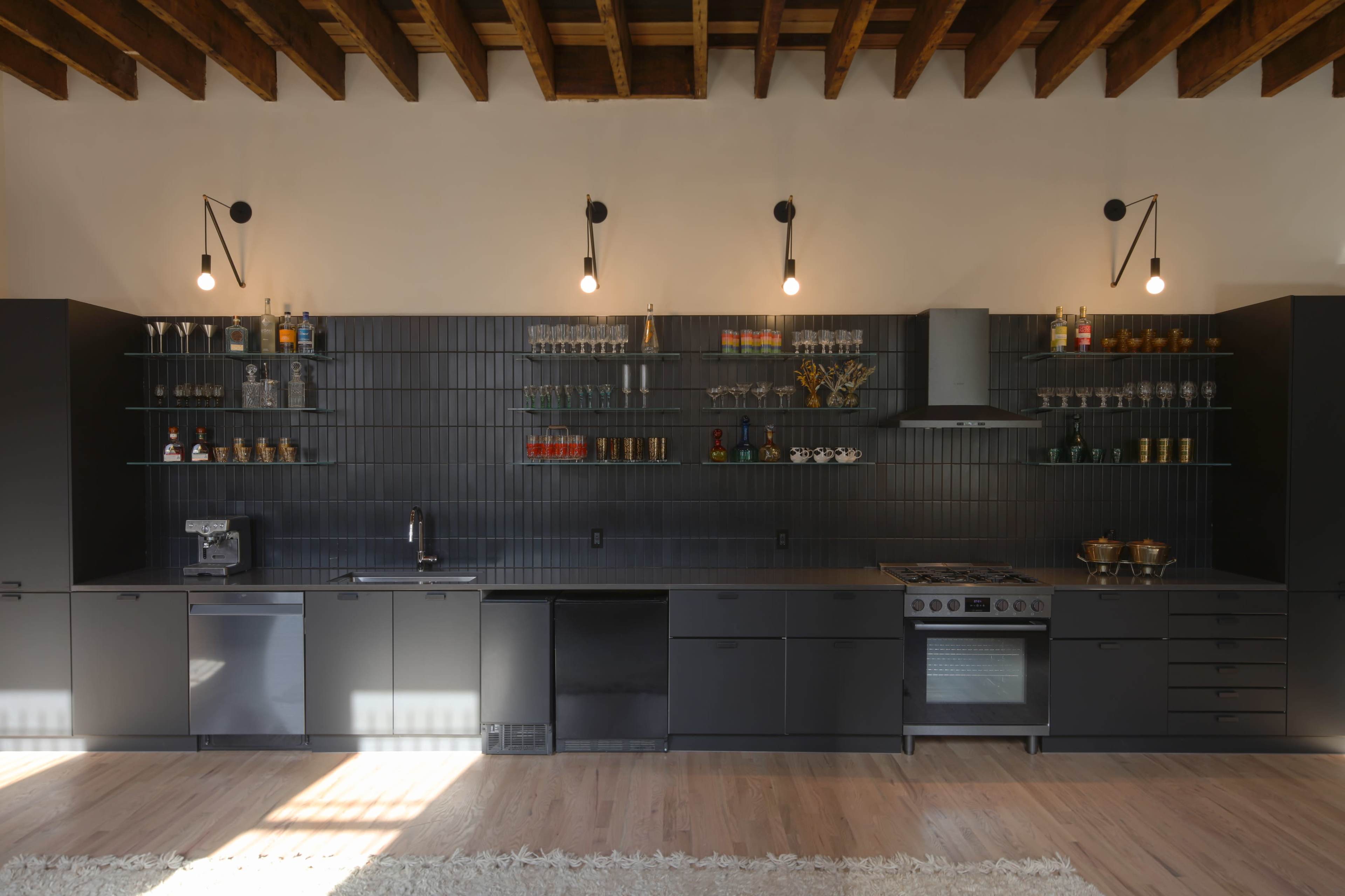 A modern kitchen with a dark themed design, including a long countertop, wall-mounted shelves holding glassware, and professional appliances.