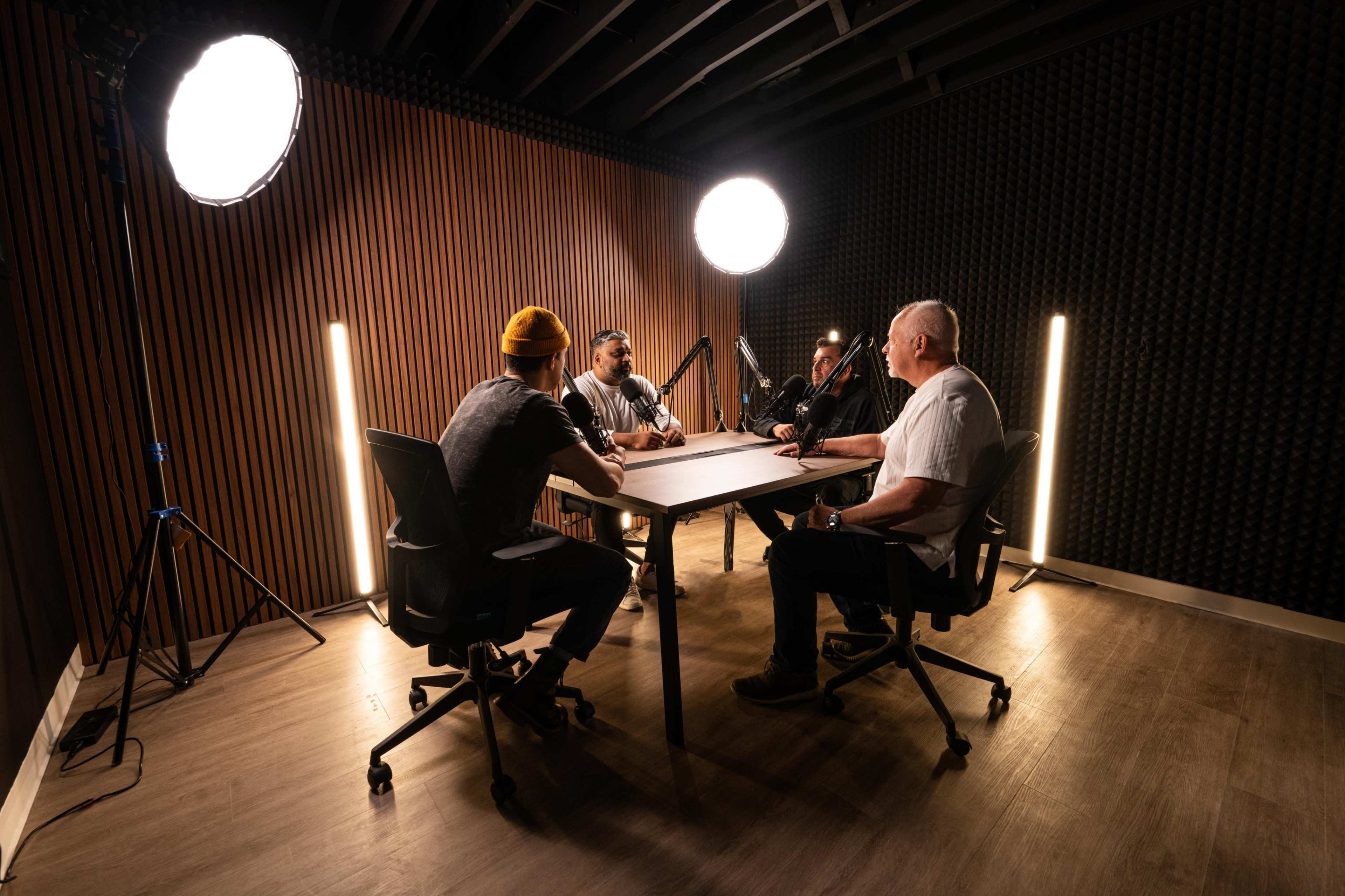 A group of five individuals is seated around a table in a podcast studio, illuminated by bright lighting, with soundproof walls in the background.