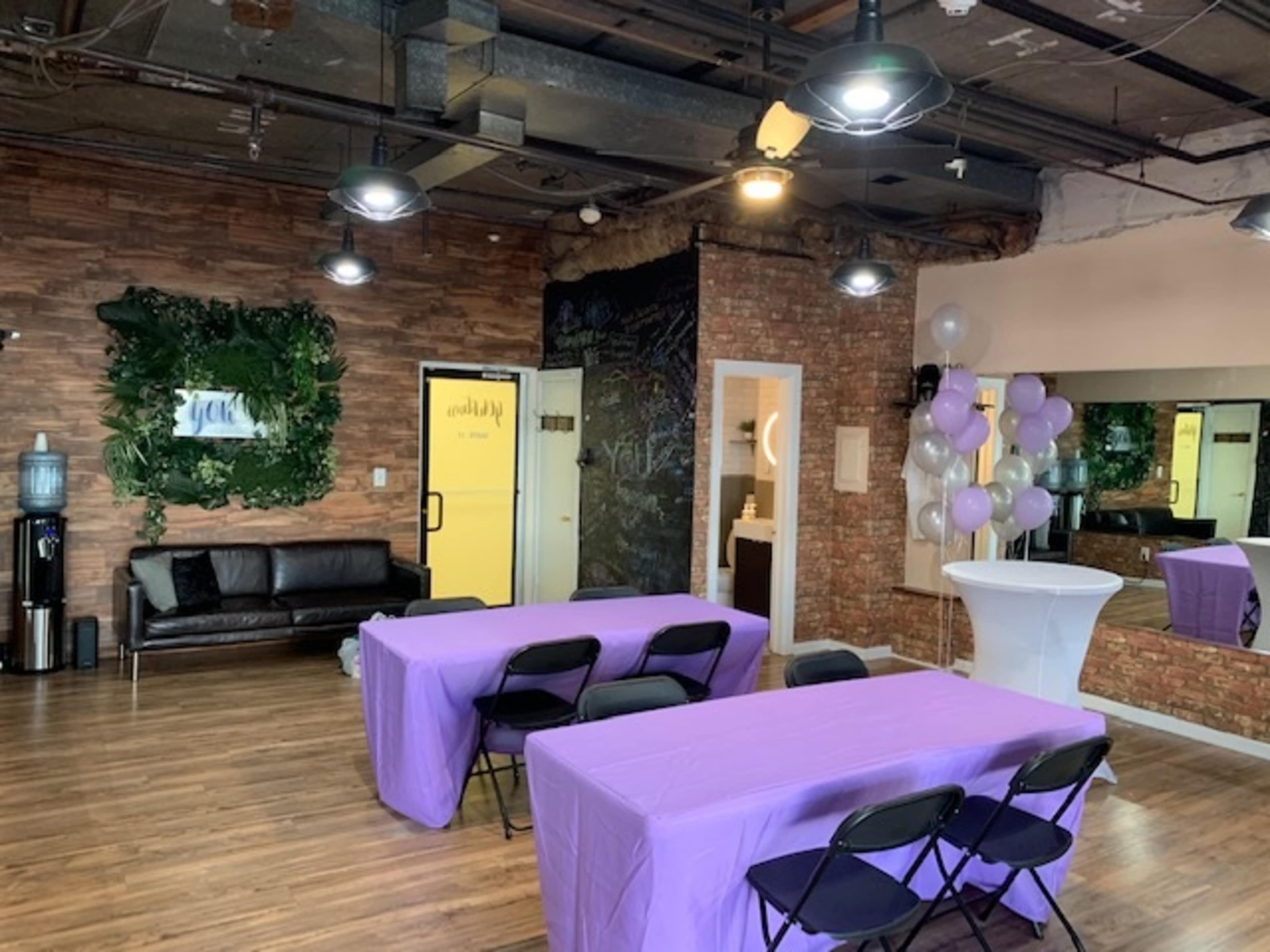 The image shows an event space with purple tablecloths, a black leather sofa, a water cooler, and a decorative wall featuring plants and a mural.