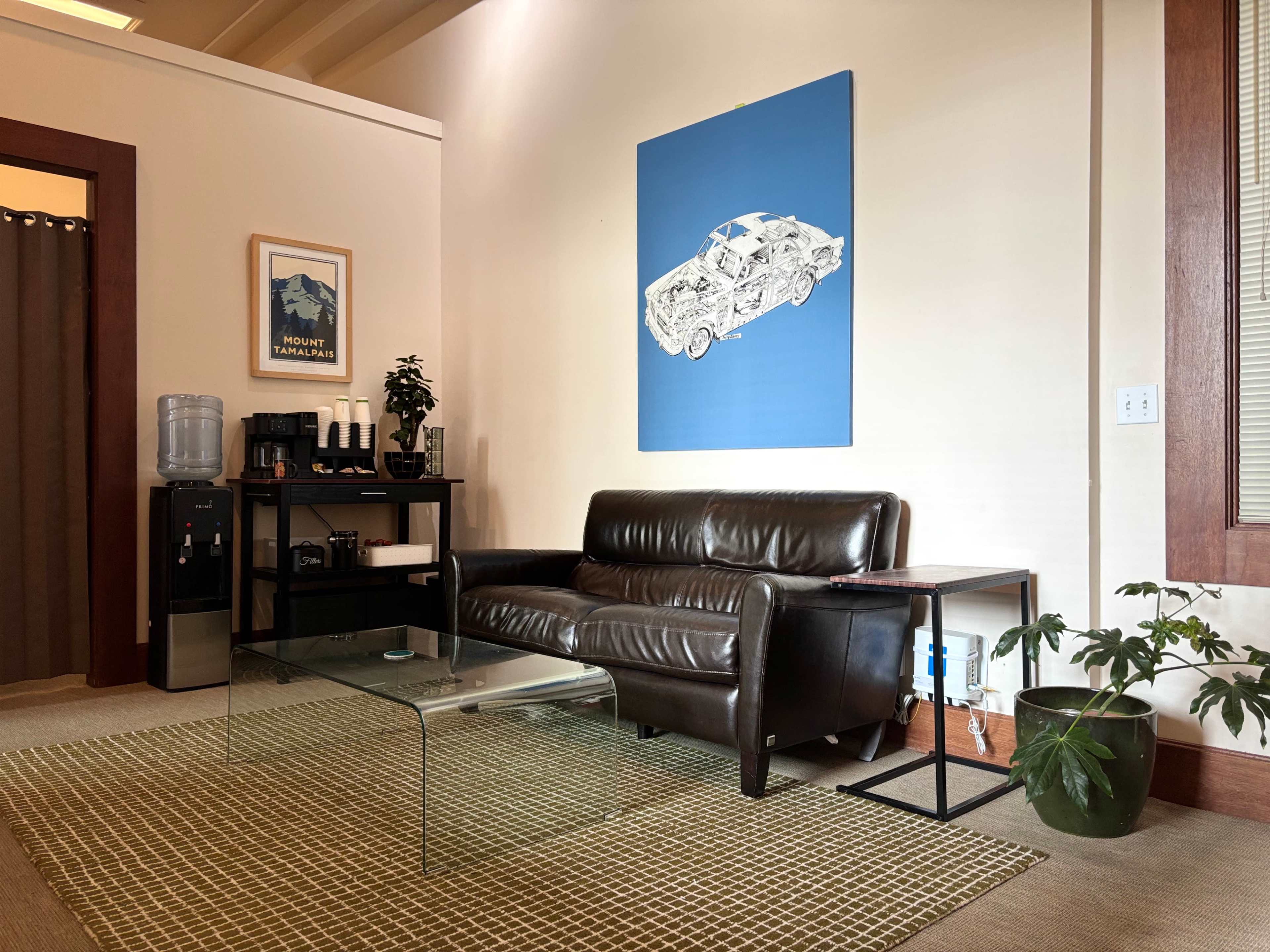 The image shows a minimalist room featuring a brown leather sofa, a glass coffee table, a water cooler, and a blue wall art piece of a car.