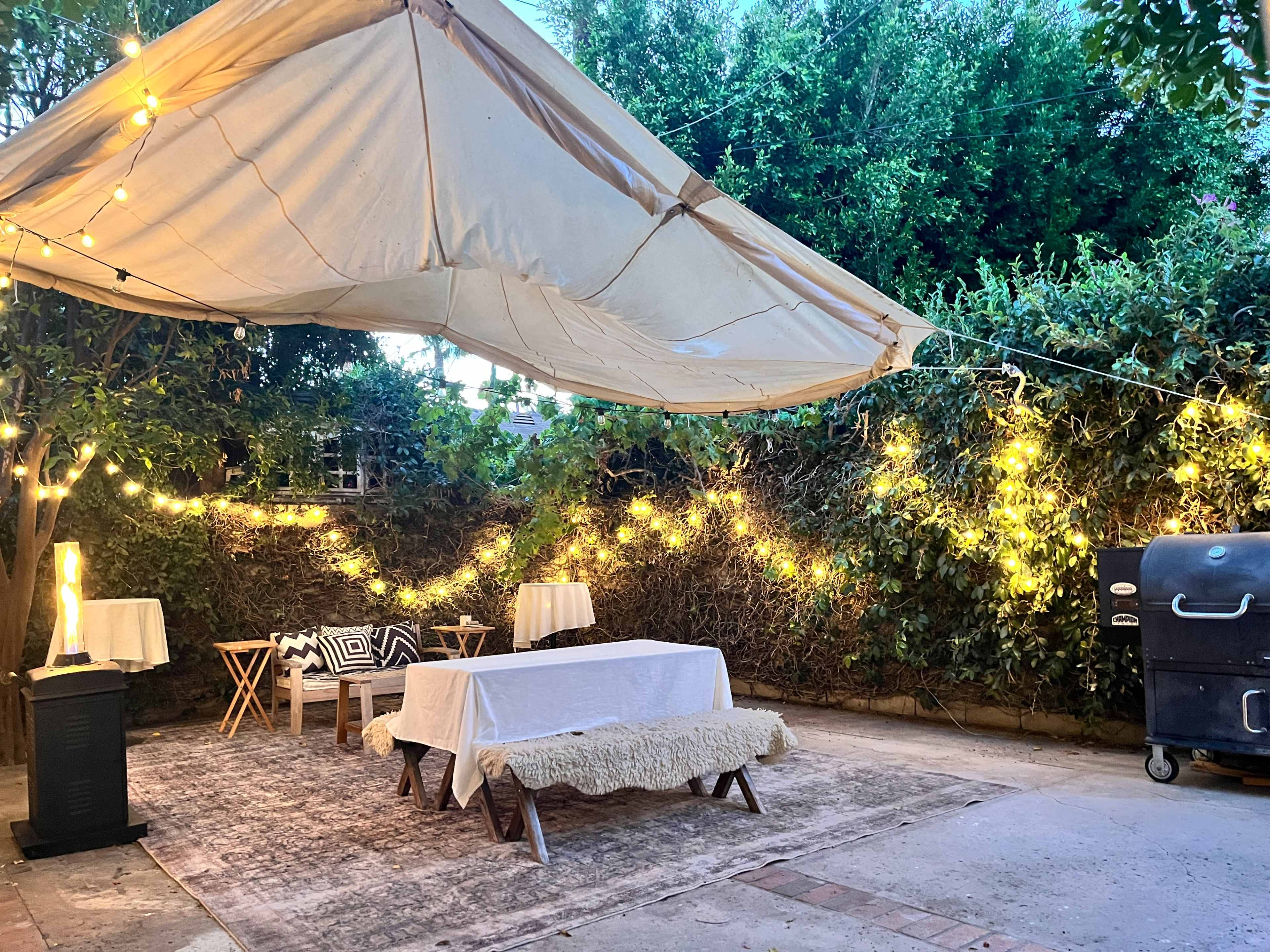 A cozy outdoor space features a large tent overhead, string lights illuminating the area, and tables arranged on a textured rug.