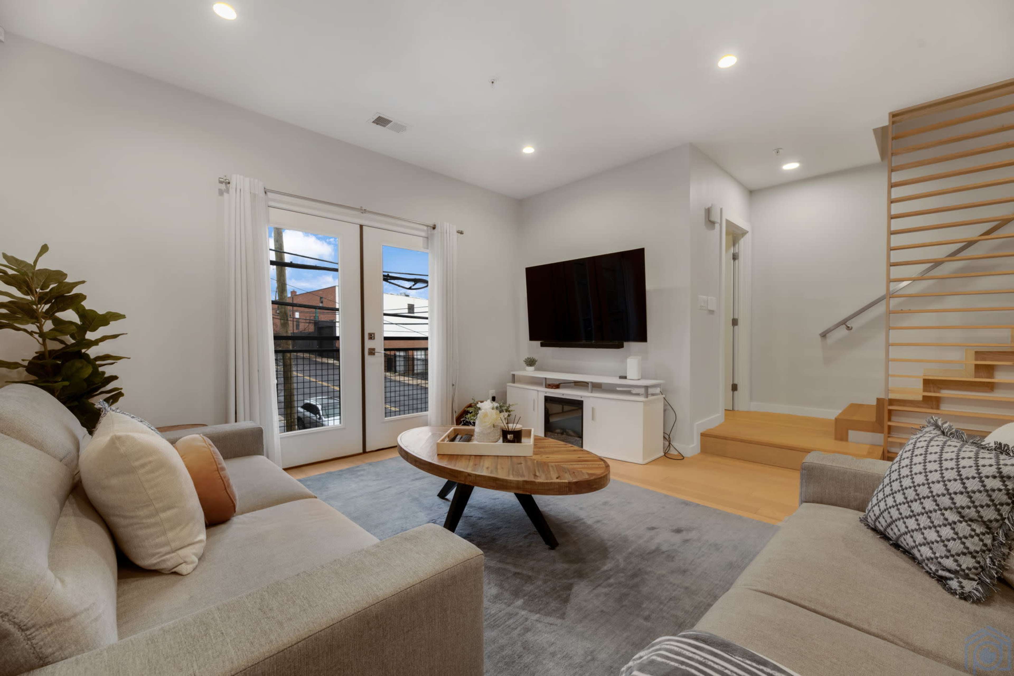 The scene features a modern living room with a light-colored sofa, a round wooden coffee table, and a large TV mounted on a white entertainment unit, alongside a staircase leading to an upper level.