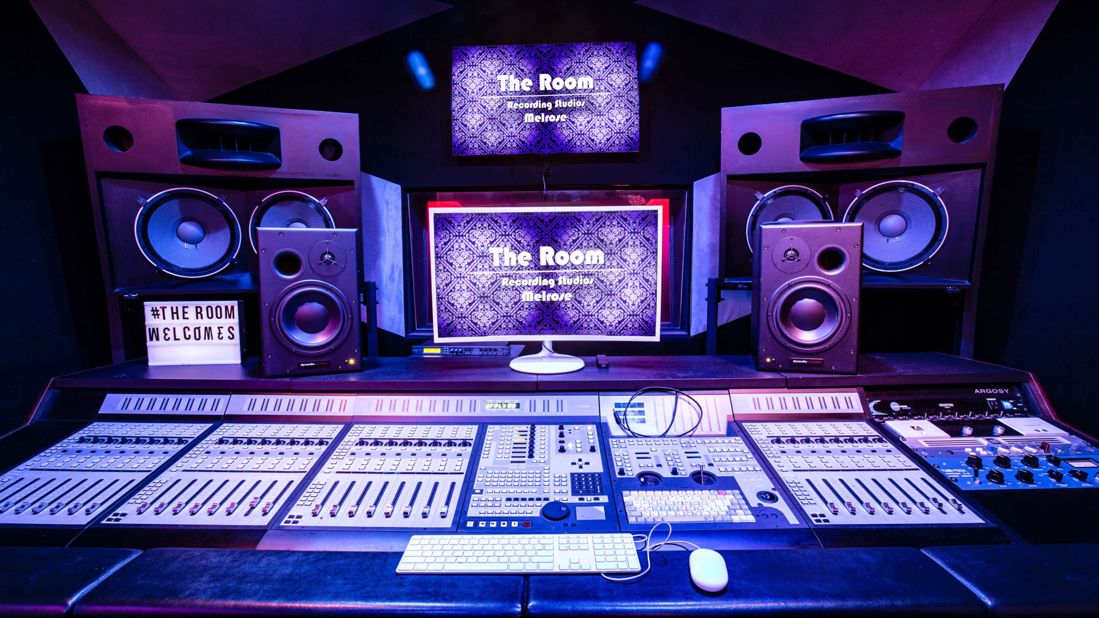The image shows a recording studio featuring a control desk with various knobs and sliders, large monitors, and two screens displaying "The Room" branding.