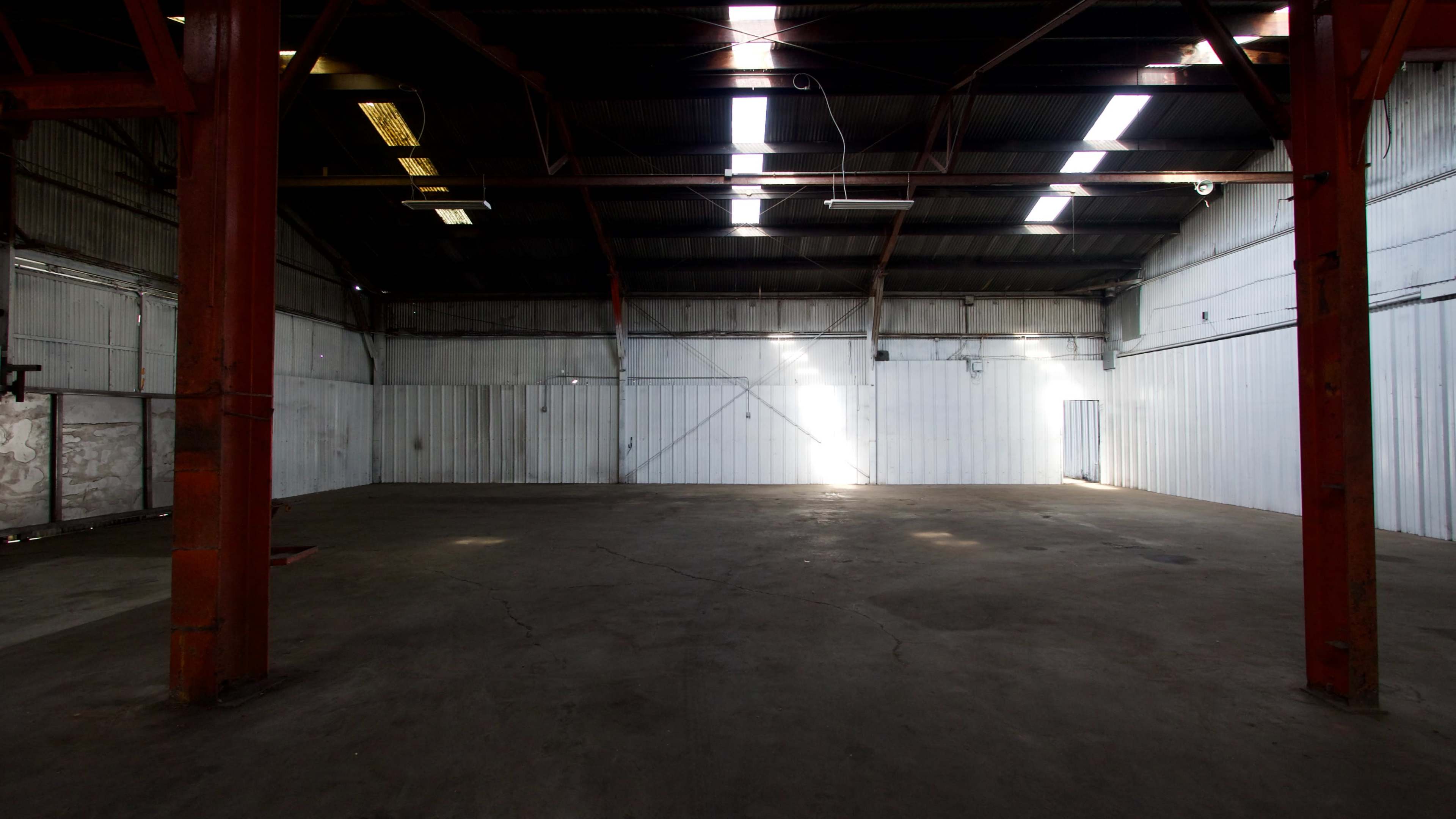 An empty industrial warehouse with concrete floors and metal beams, illuminated by natural light coming through skylights.