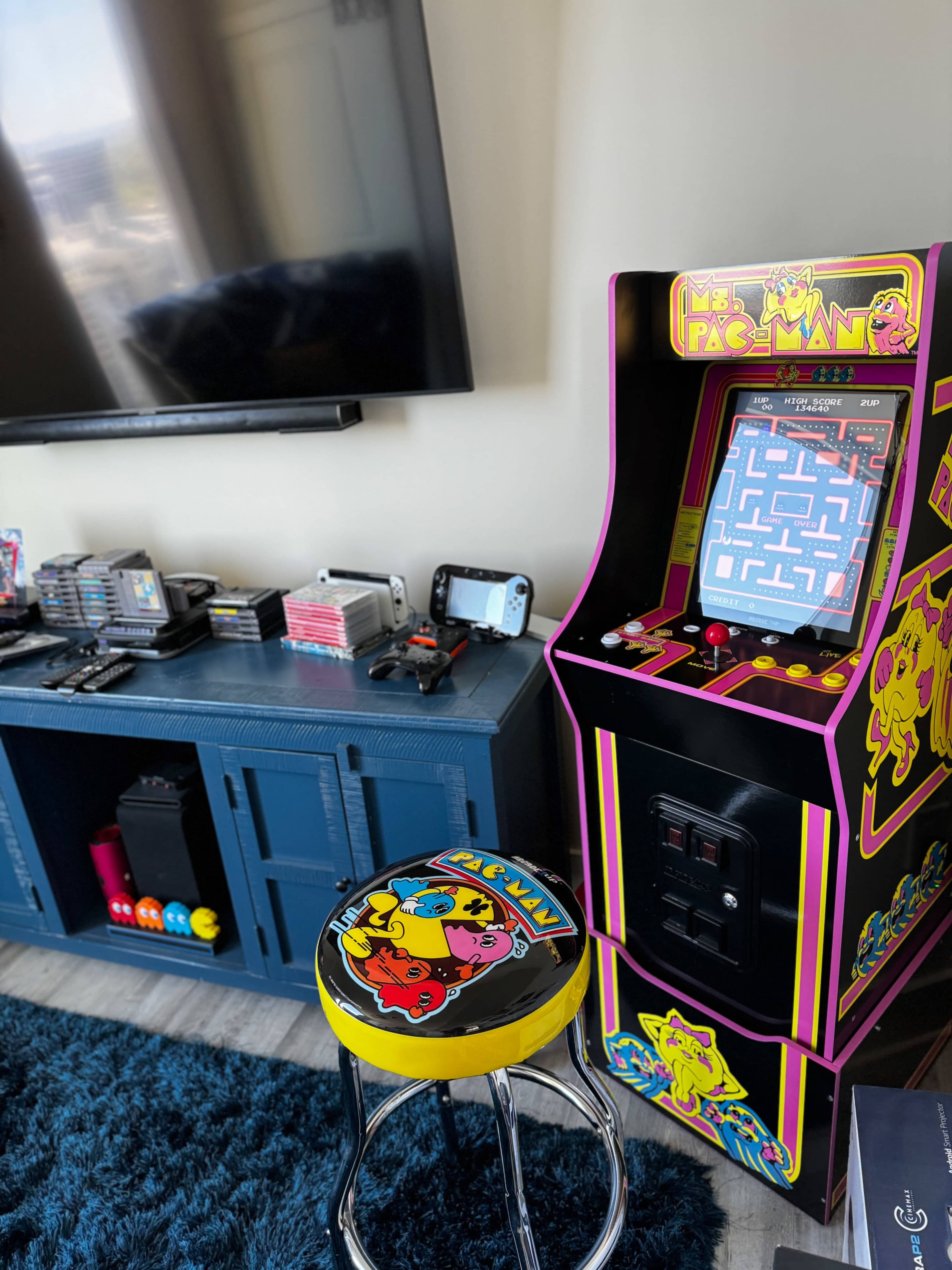 A colorful arcade cabinet featuring "Ms. Pac-Man" stands next to a blue console table with various gaming consoles and accessories in a living room setting.