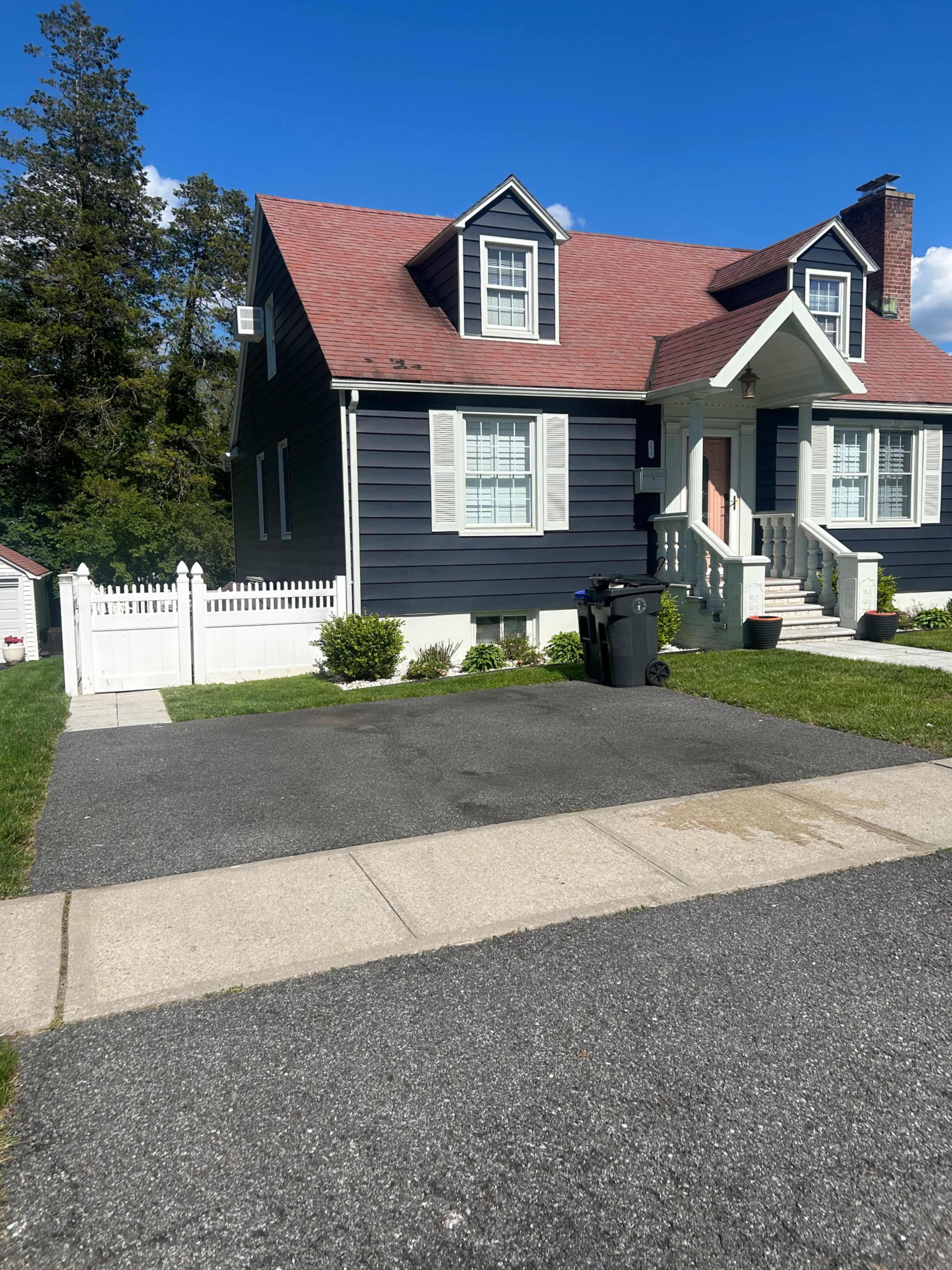 A two-story house with dark blue siding and a red roof has a white picket fence and a driveway leading to the front entrance.