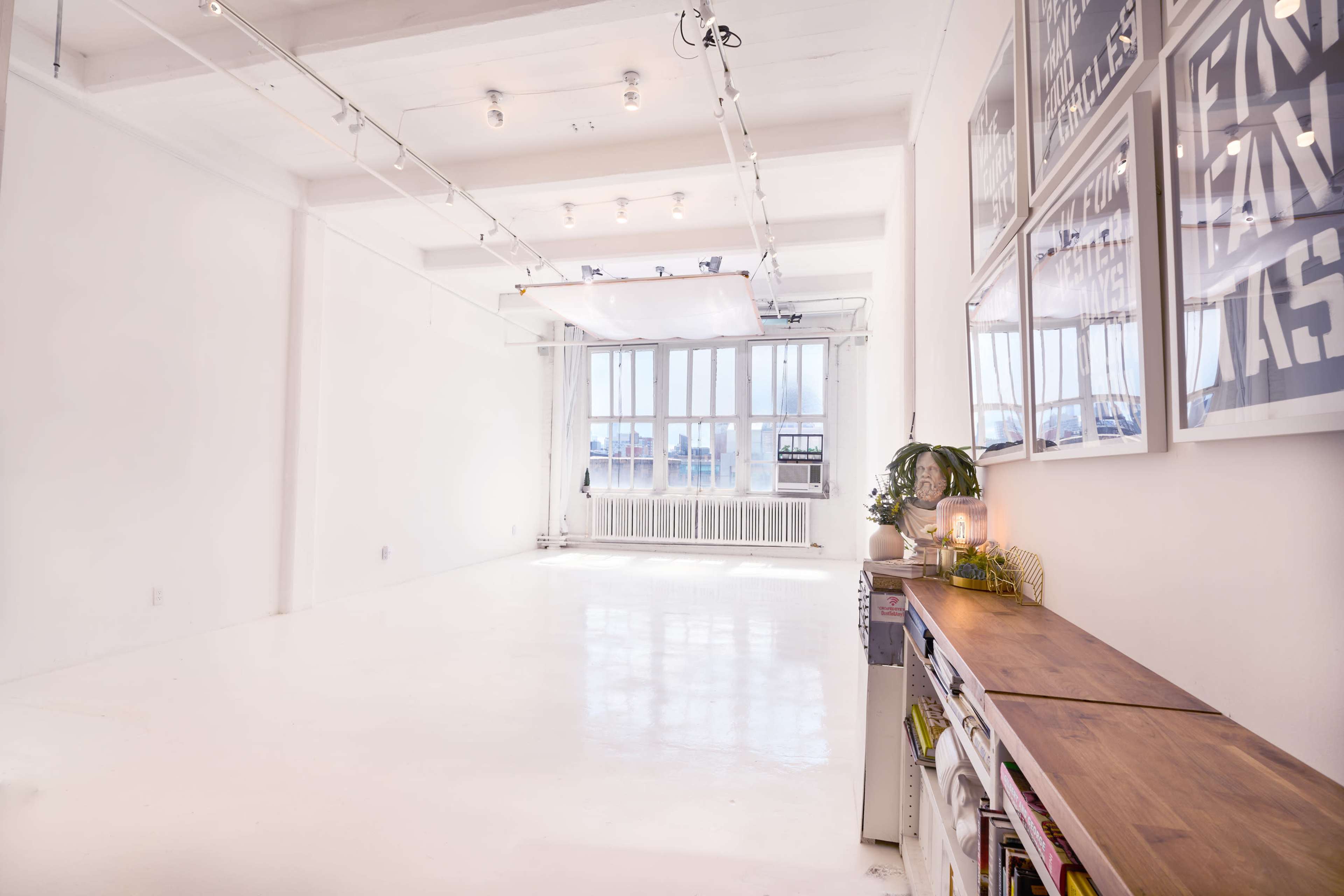 A spacious, well-lit studio features white walls and floors, with large windows and a wooden shelf against one side.