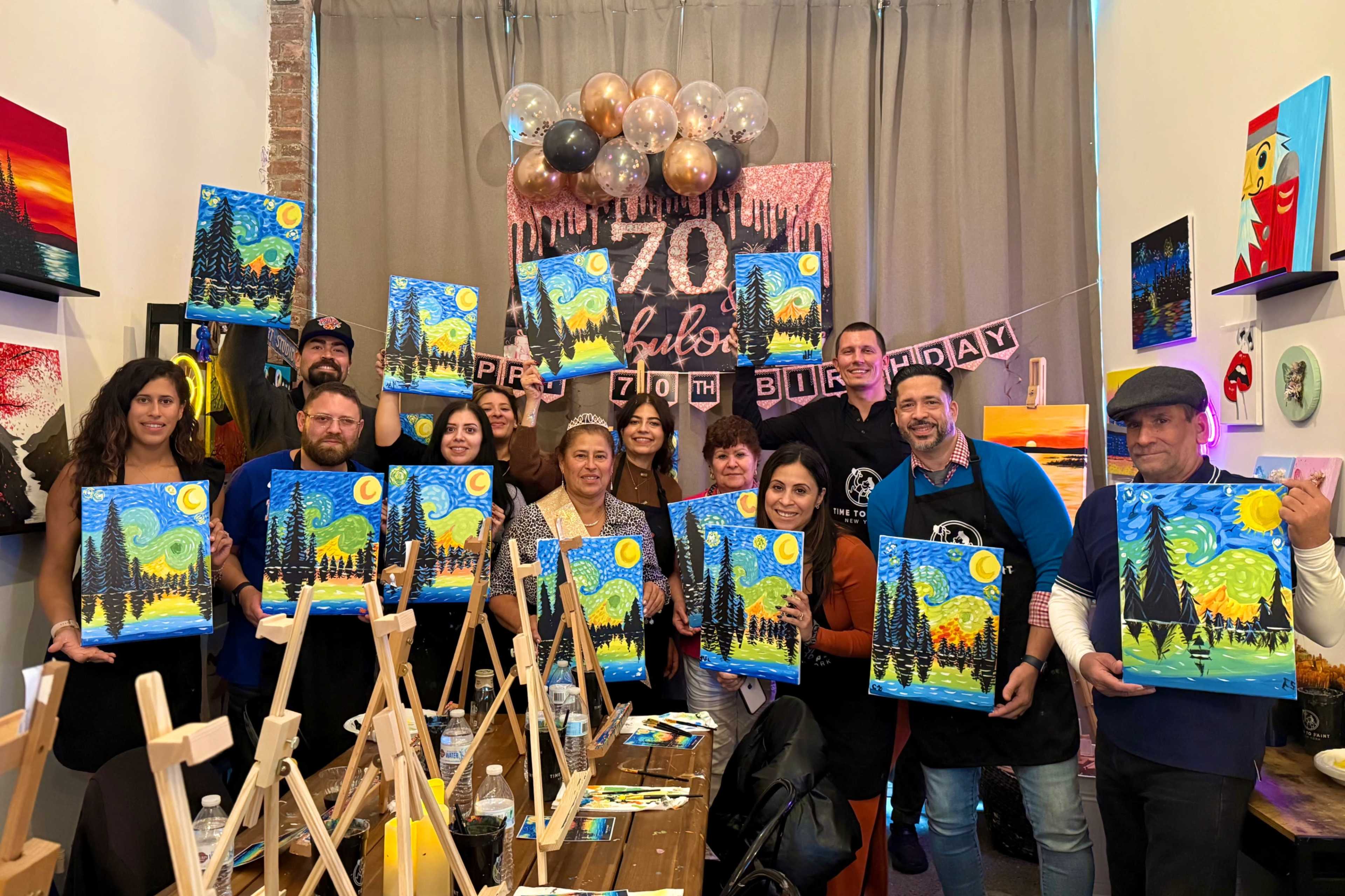 A group of people holds up their completed paintings during a birthday celebration inside a decorated room.