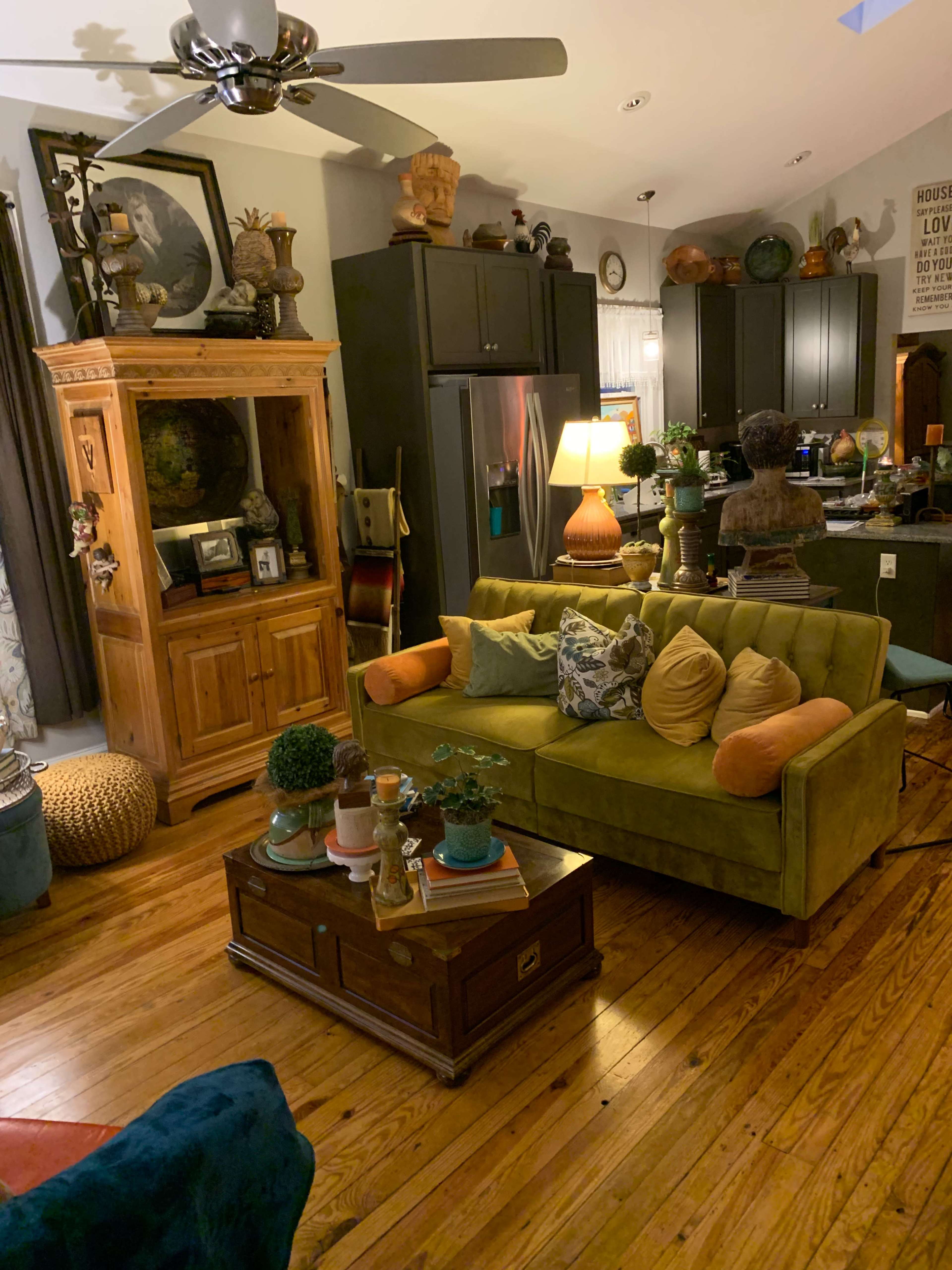 A cozy living room with a green sofa, wooden furniture, decorative plants, and a well-lit kitchen area in the background.