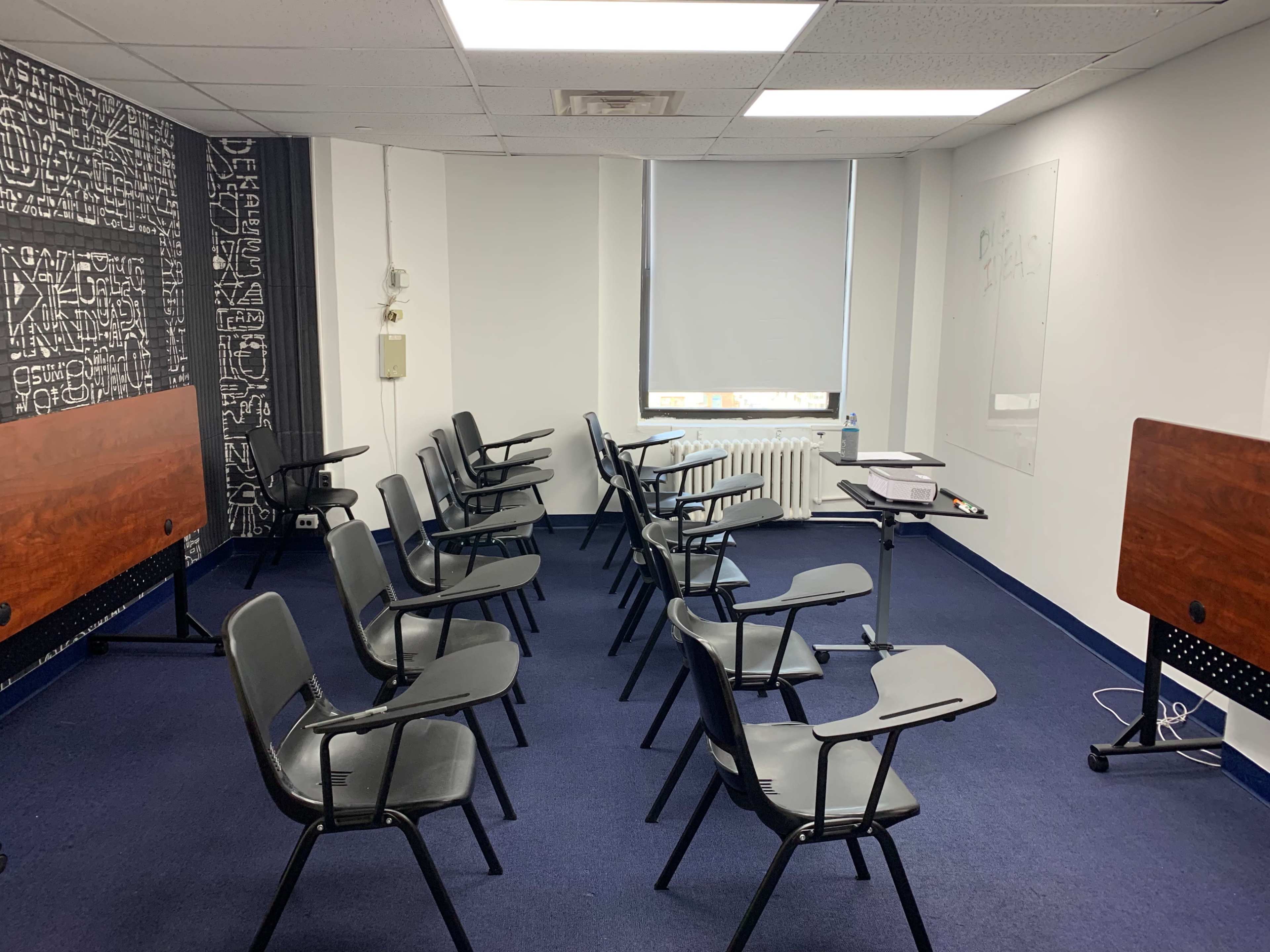 A classroom setting features several rows of black chairs with attached writing surfaces, arranged facing a large whiteboard and a wall covered in abstract designs.