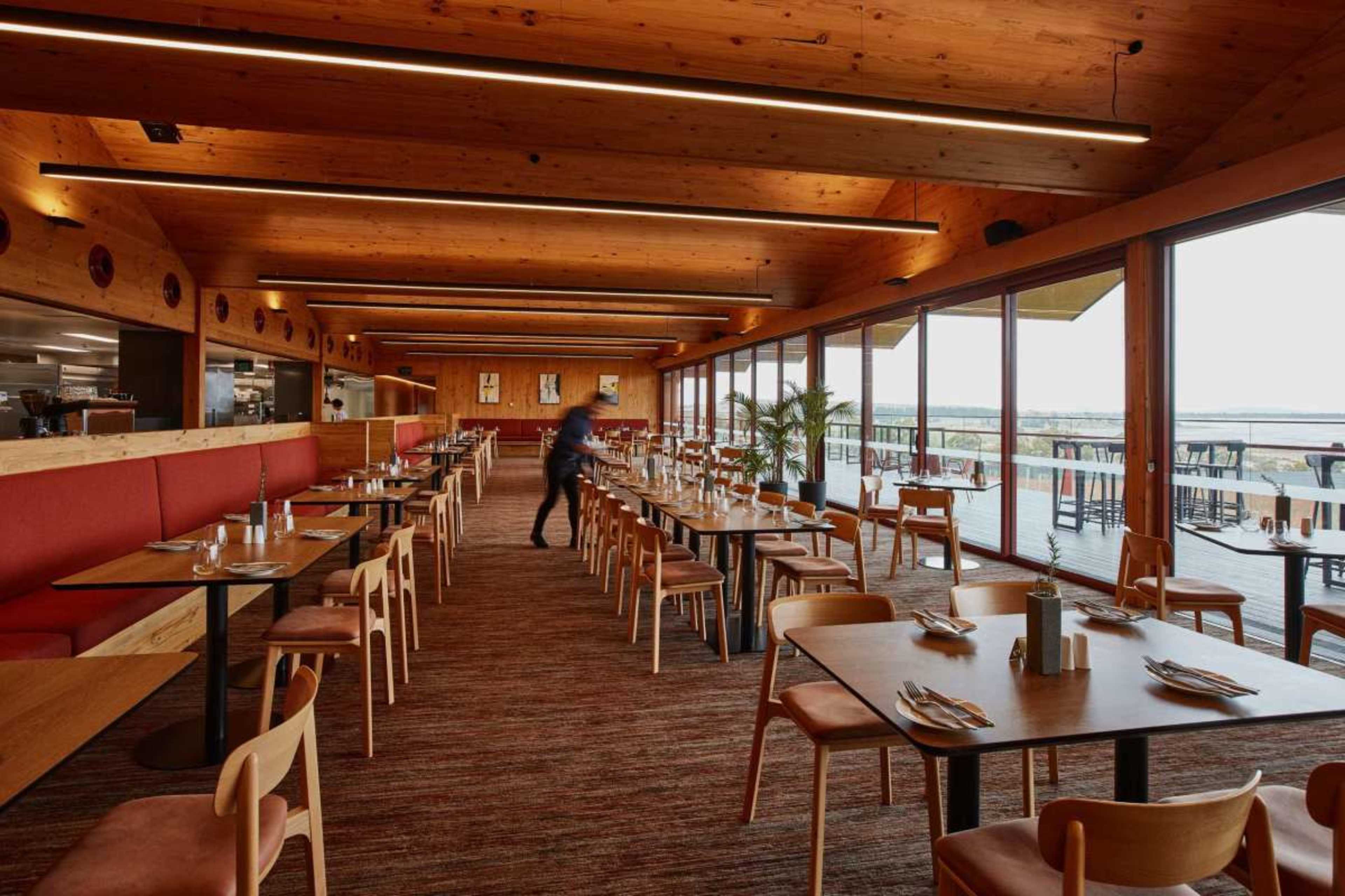 The image shows a modern restaurant interior featuring wooden beams, large windows with views outside, and rows of tables and chairs set for dining.