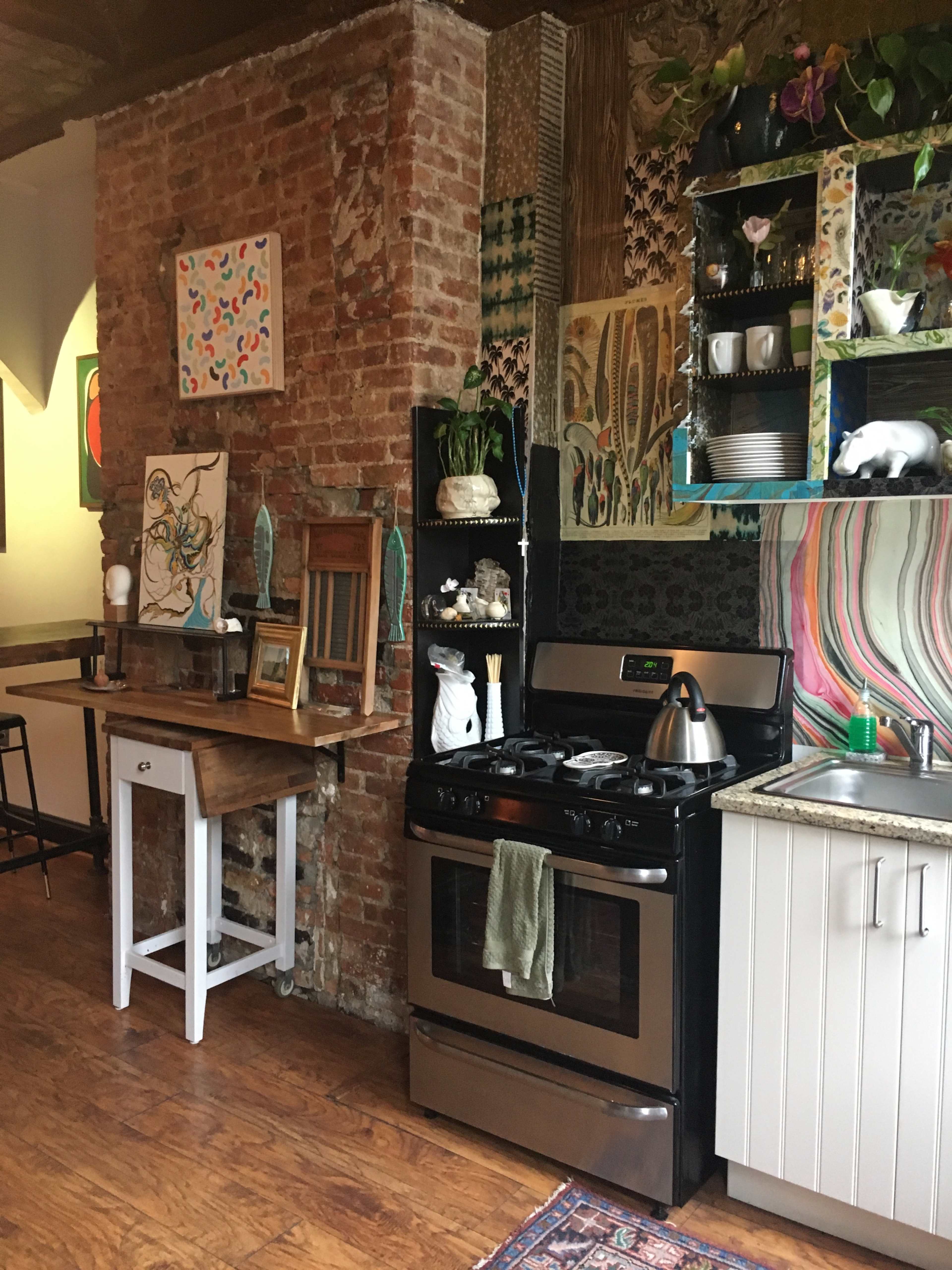 A rustic kitchen with a brick wall, a stainless steel stove, a wooden table, and colorful decor along the walls and shelves.