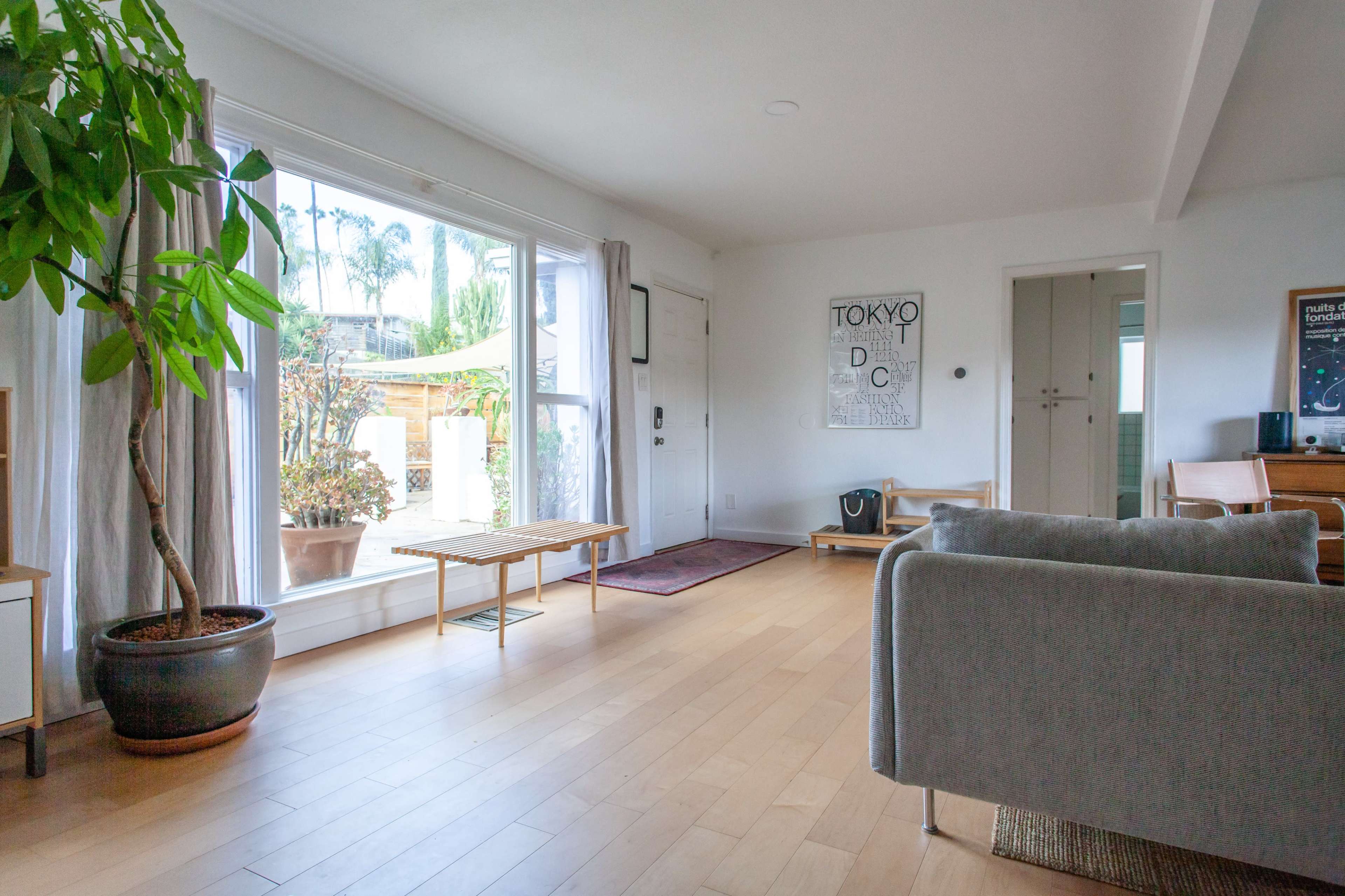 A bright living room with wooden flooring, large windows, and minimalist furniture including a grey sofa and a small wooden bench.