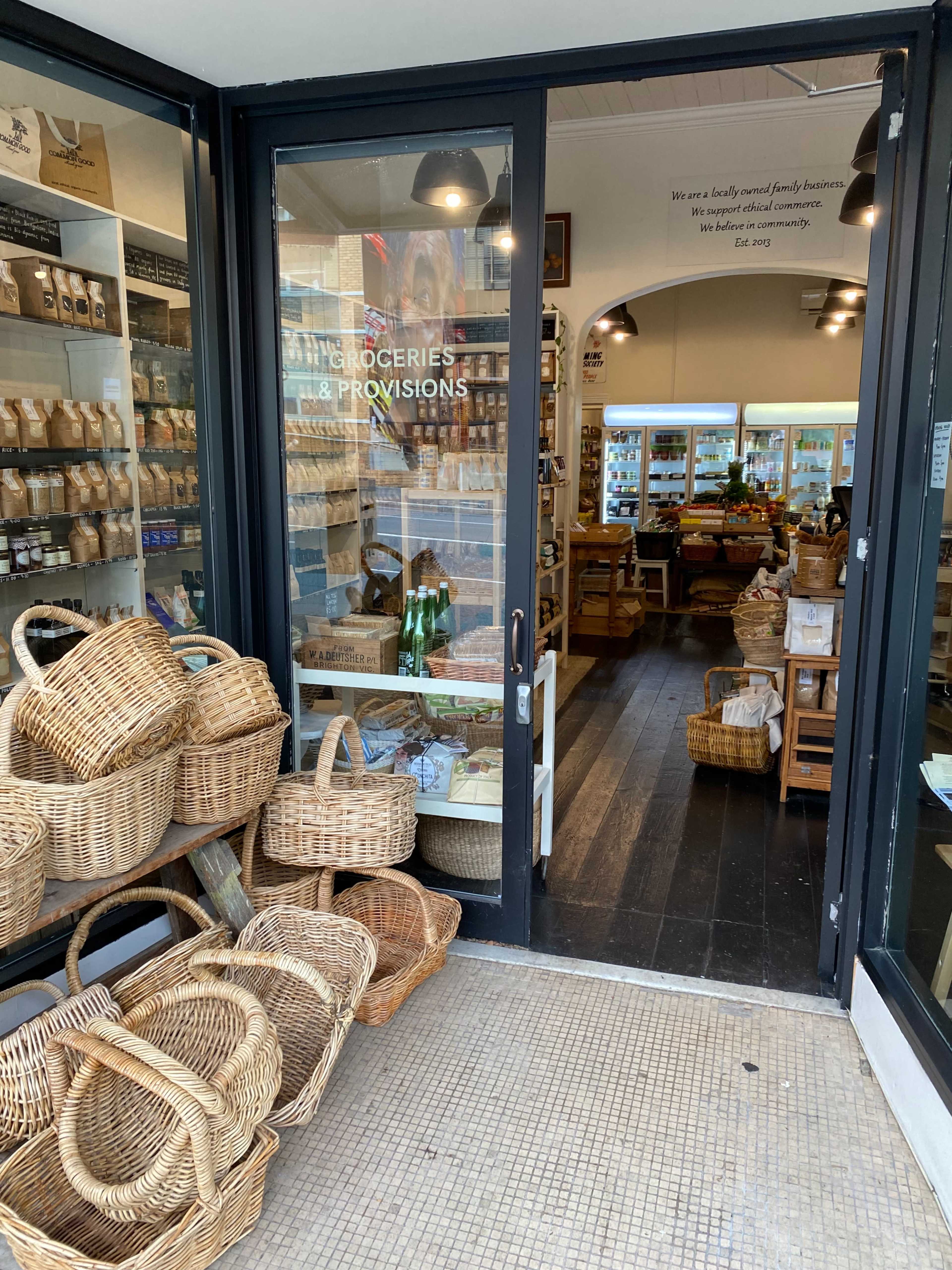 A storefront displays a variety of grocery items and provisions, with woven baskets arranged outside the entrance.